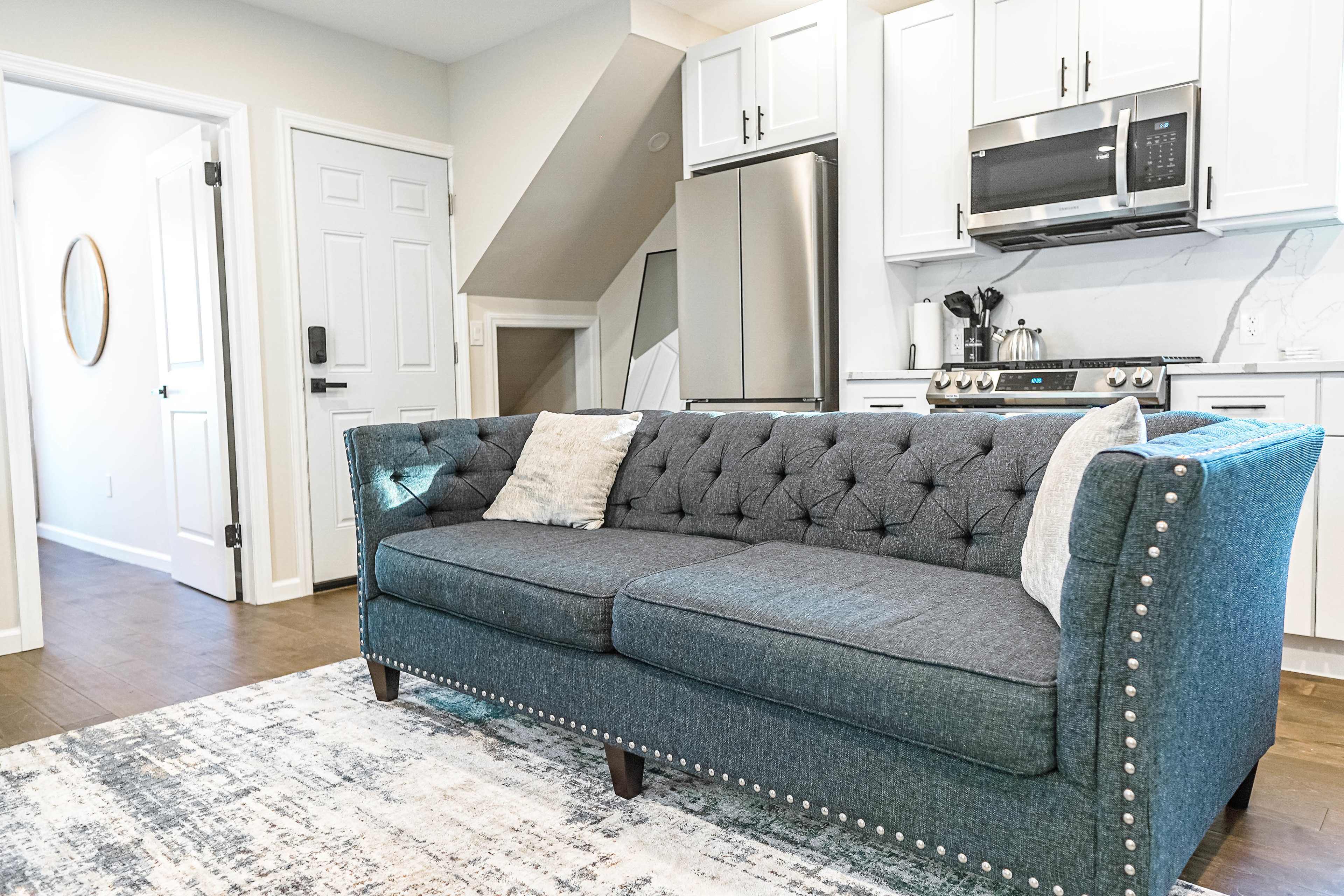 A modern, tufted teal sofa placed in a well-lit kitchen area that features stainless steel appliances and white cabinetry.