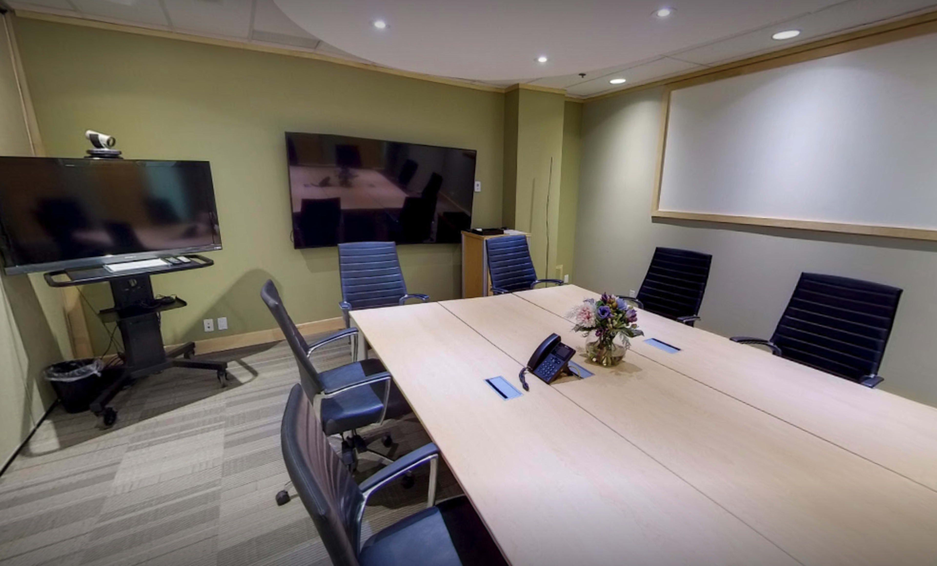 A conference room features a large table surrounded by black chairs, with a television and whiteboard mounted on the walls.