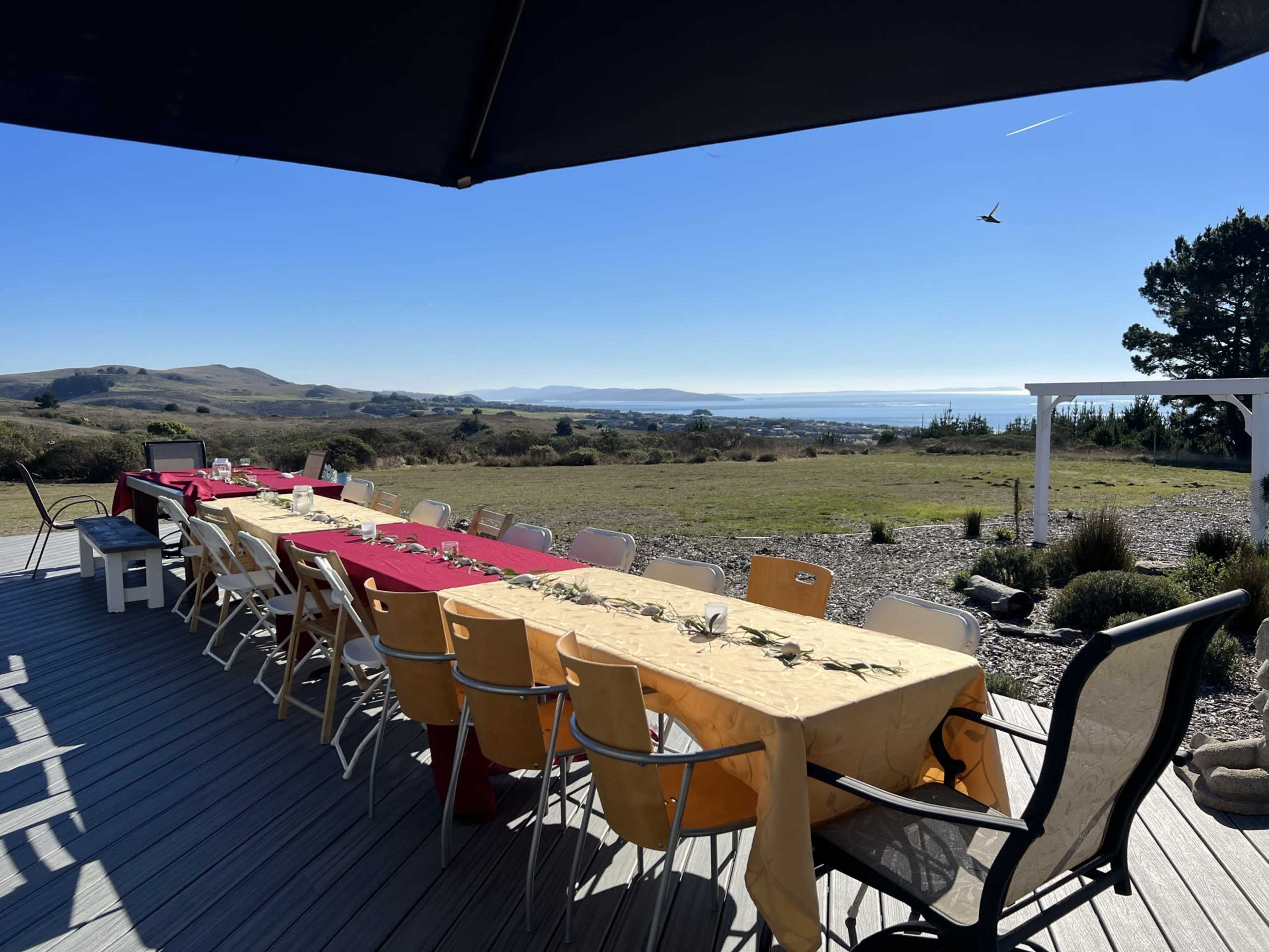 A long outdoor table is set with colorful tablecloths, surrounded by chairs, overlooking a scenic view of hills and a body of water.