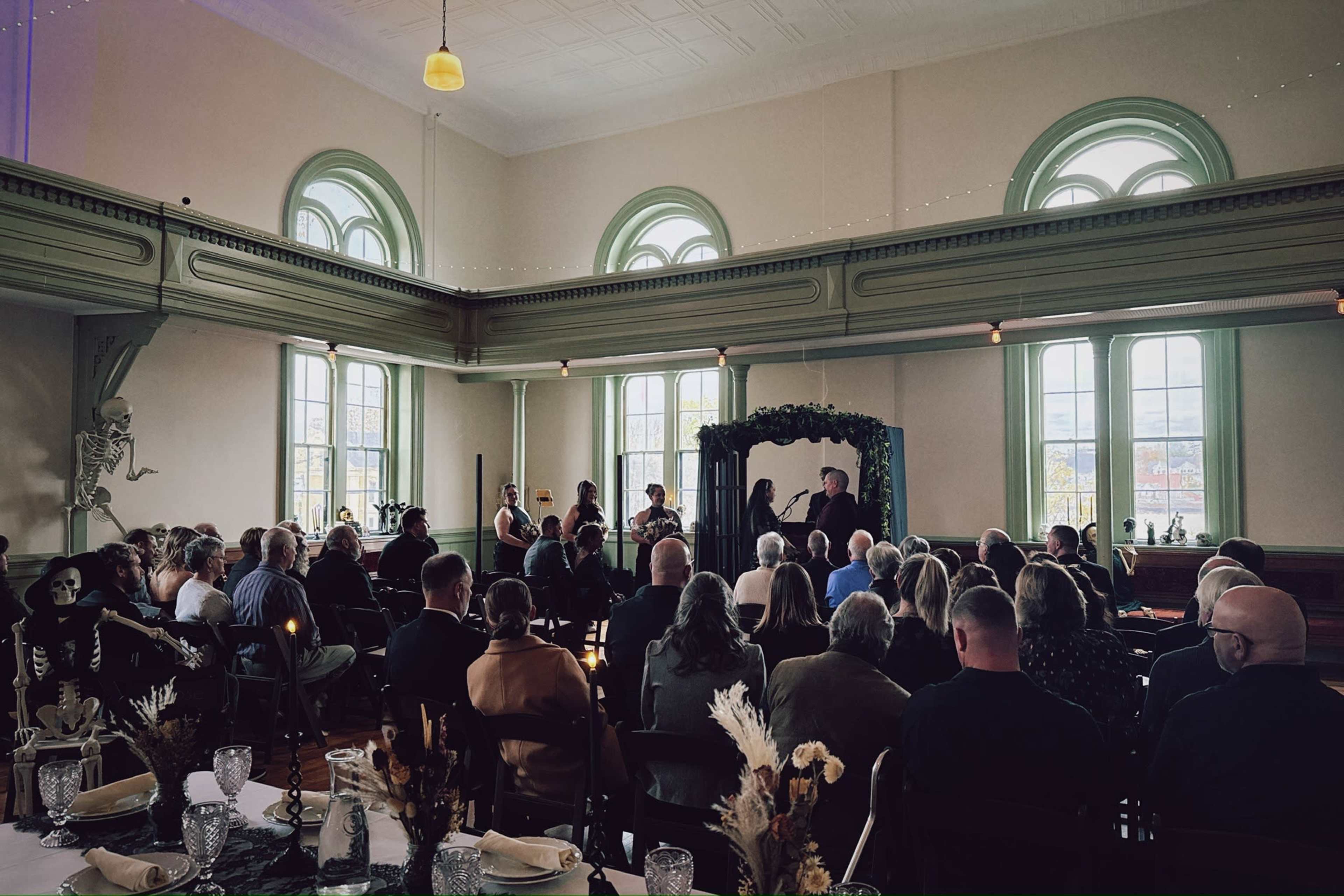 A crowd gathers inside a well-lit venue with large windows, as a small group performs near a decorative arch.