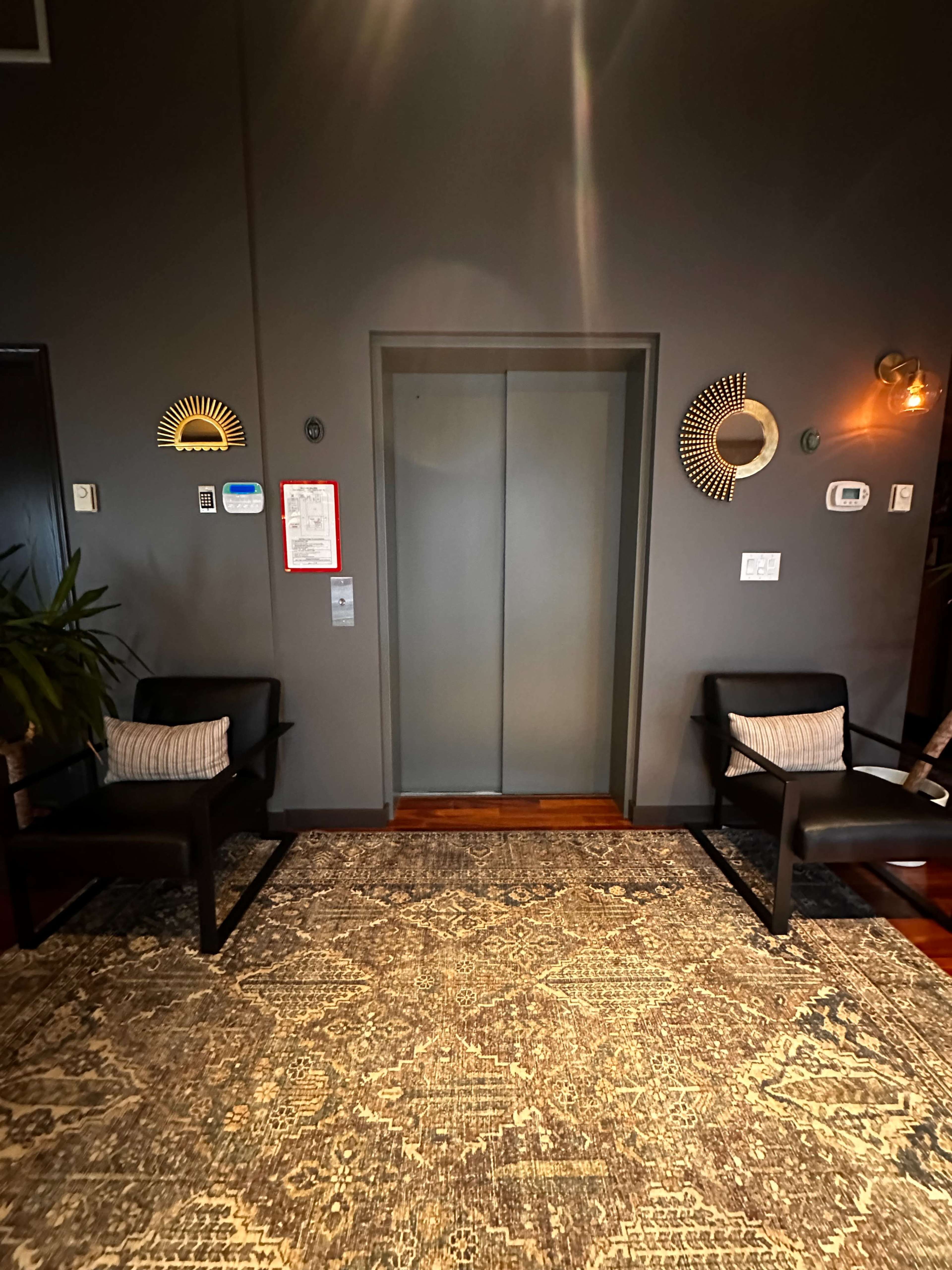 The image shows a hallway with an elevator door in the center, flanked by two black chairs and a decorative rug on the floor.