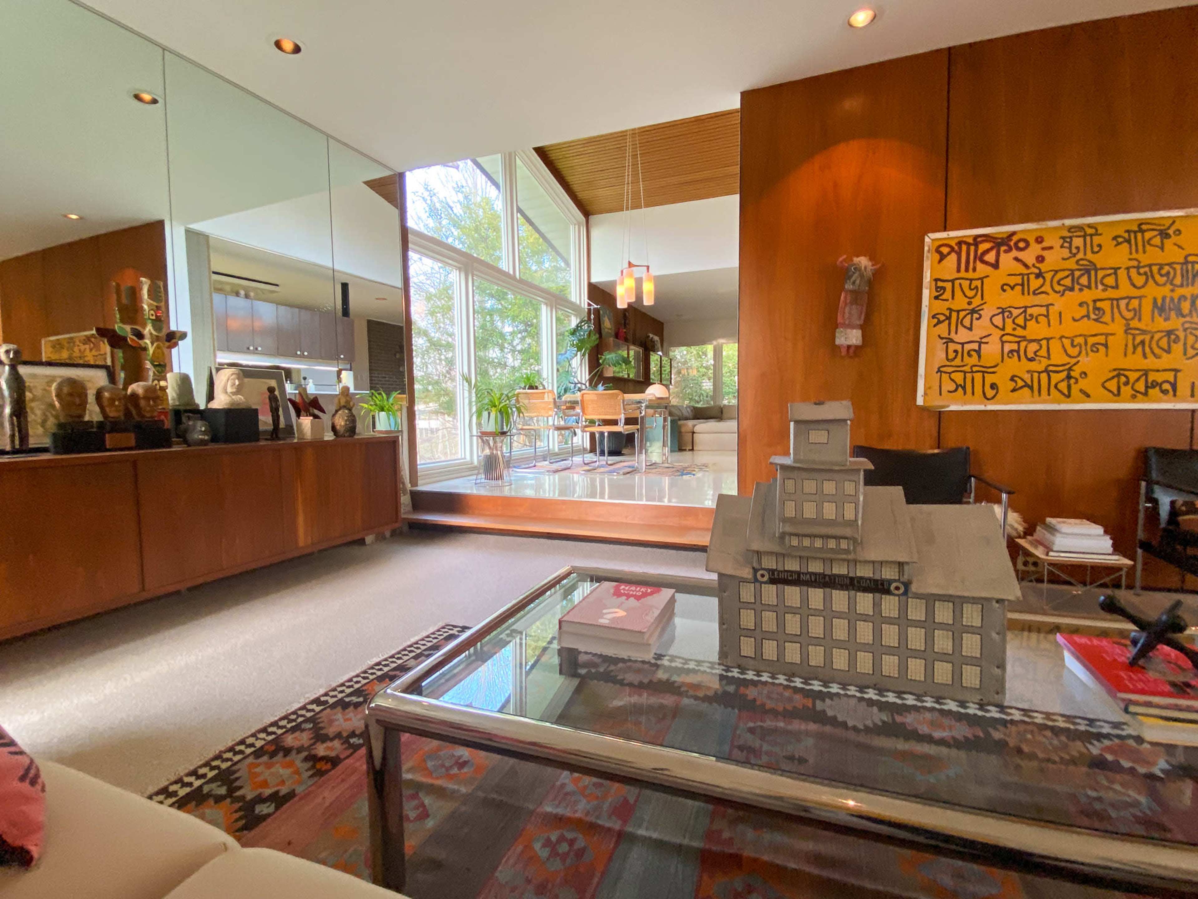 The image features a modern interior space with a wooden accent wall, large windows letting in natural light, and various decorative elements including sculptures and a model building on a coffee table.