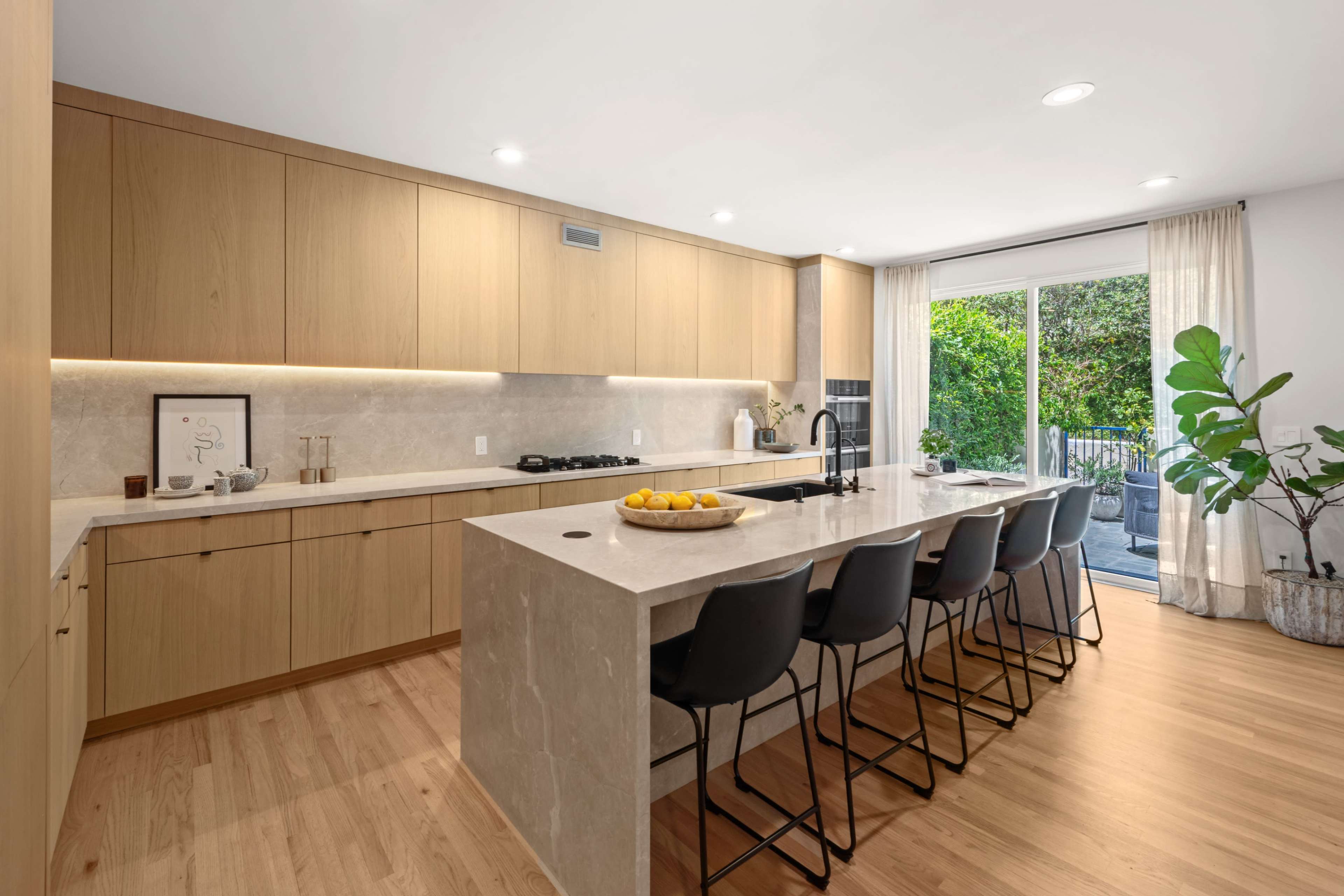 The modern kitchen features wooden cabinetry, a large island with seating, and a sliding door that opens to an outdoor area.
