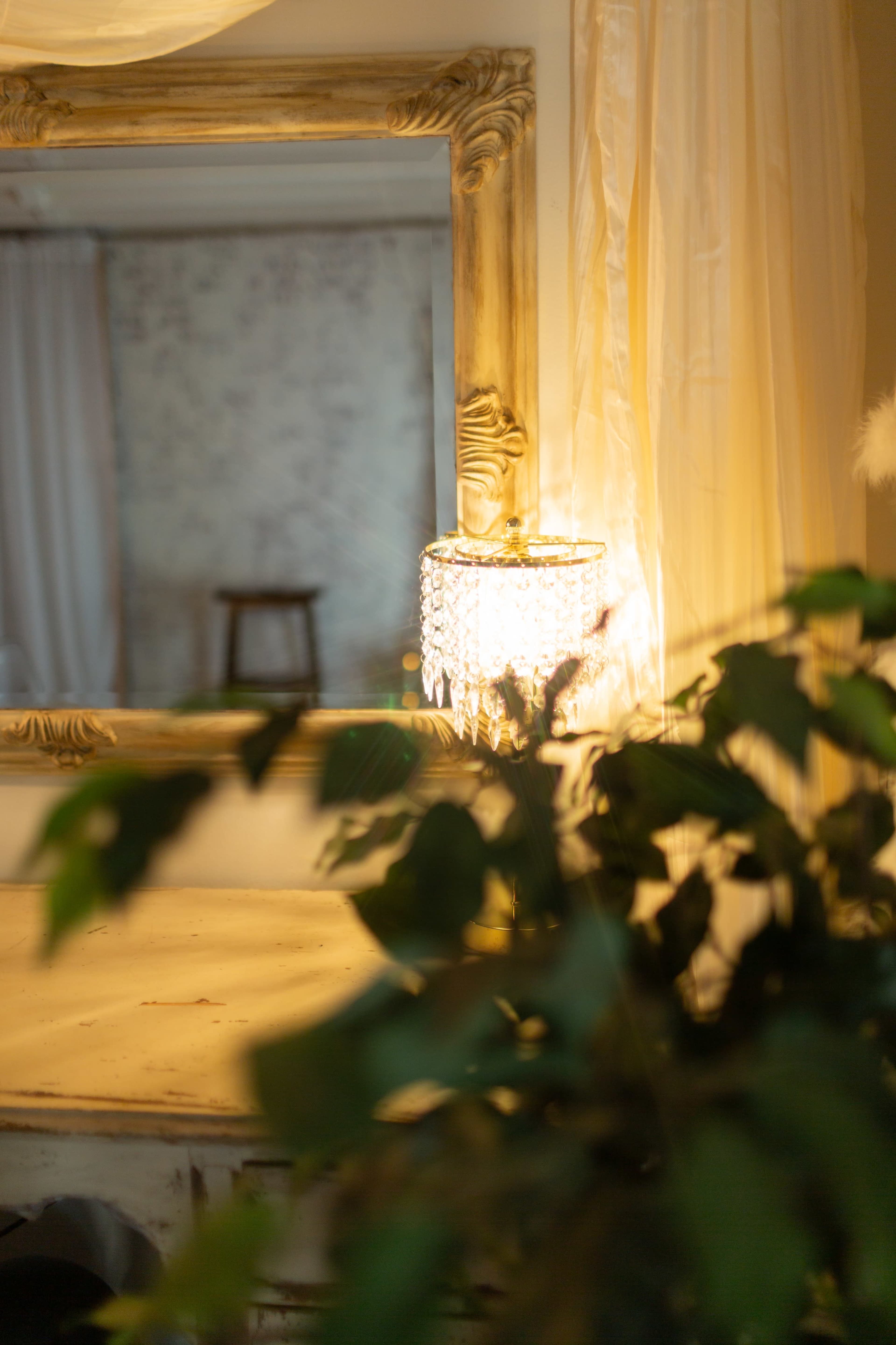 The image features a decorative mirror framed in an ornate design, beside a shimmering lamp, with a lush greenery in the foreground.