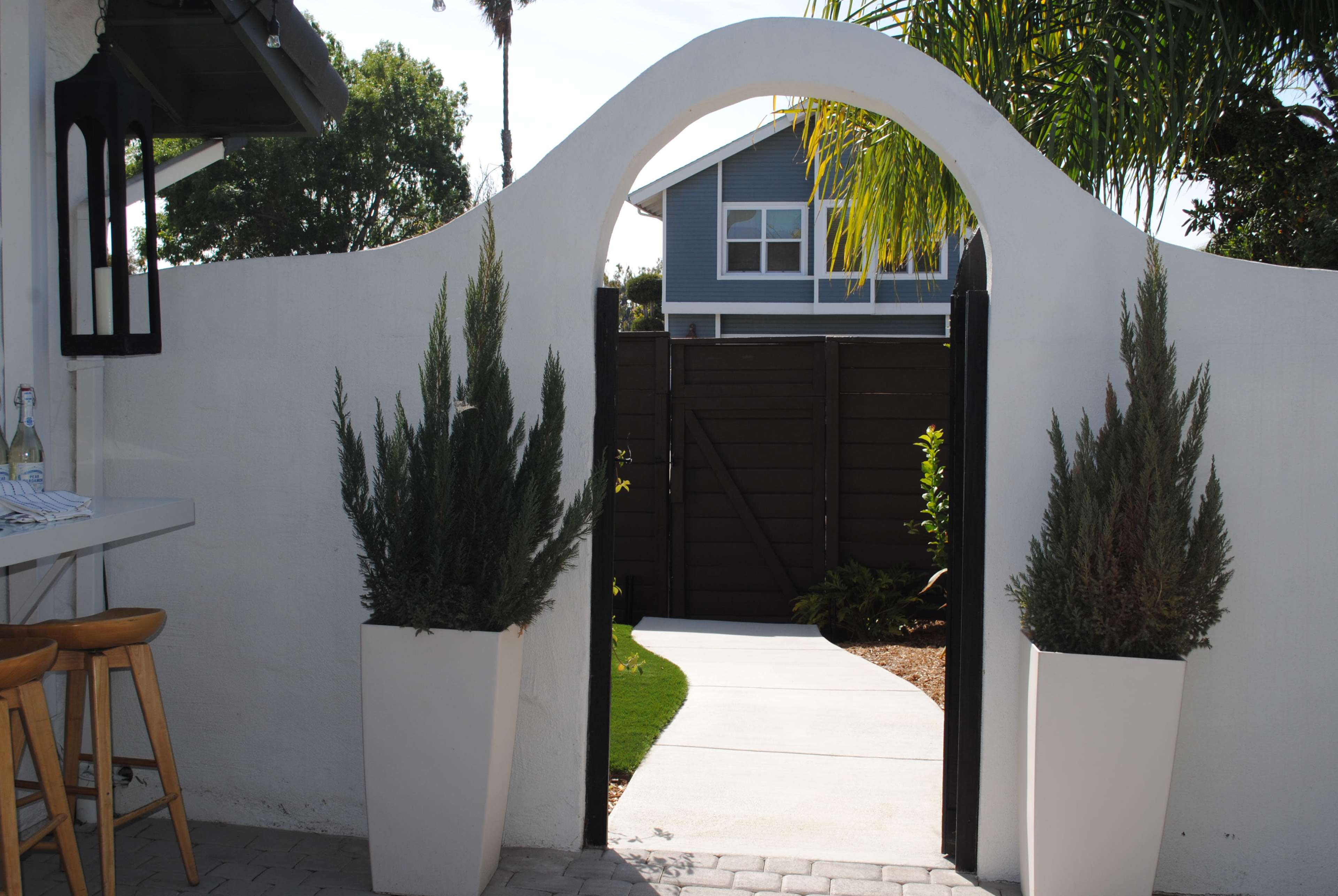 A white archway frames a path leading to a fenced garden area, with potted plants on either side.