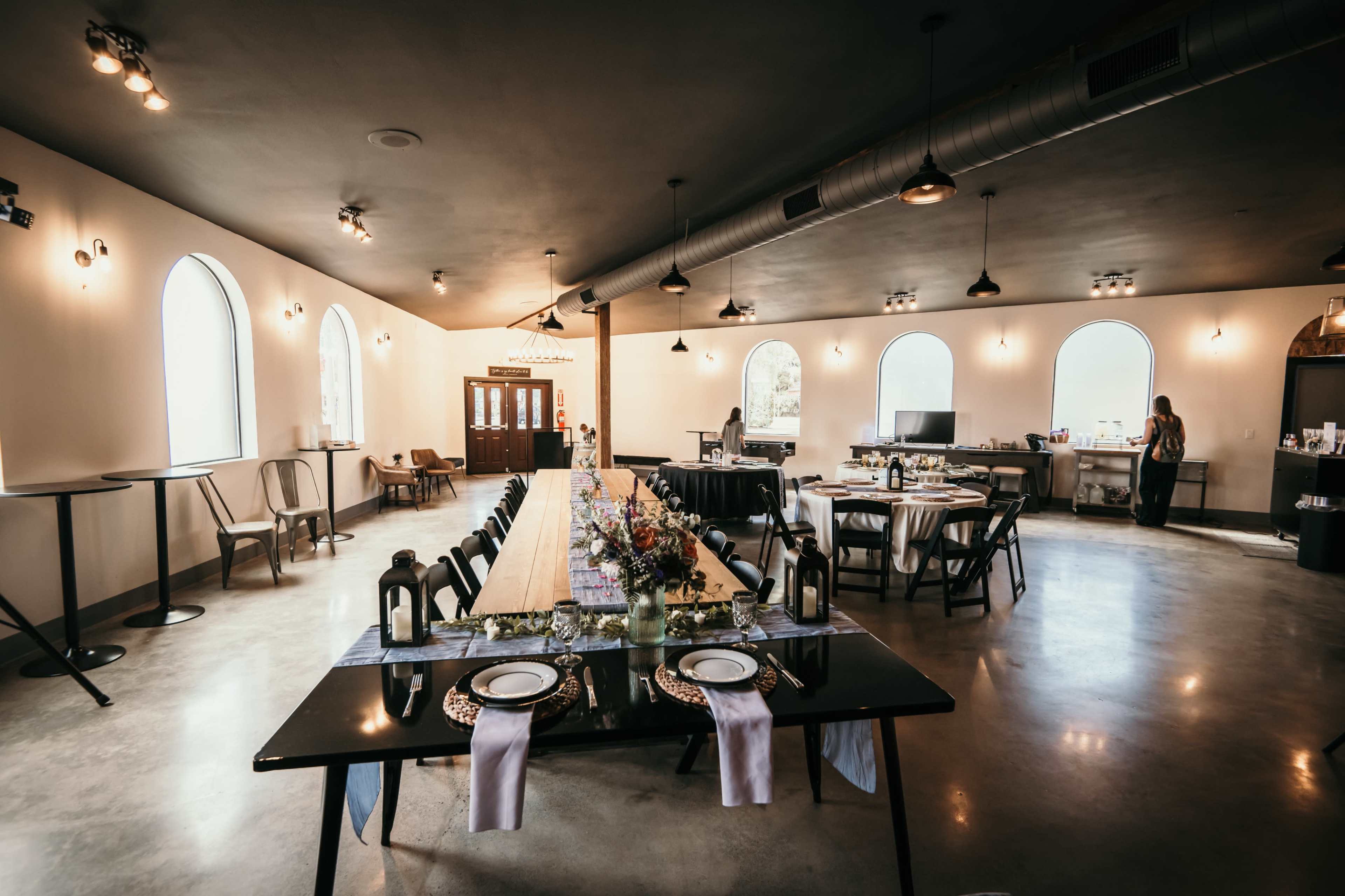 A spacious event room features a long central table set for dining, surrounded by additional tables and chairs, with large arched windows letting in natural light.