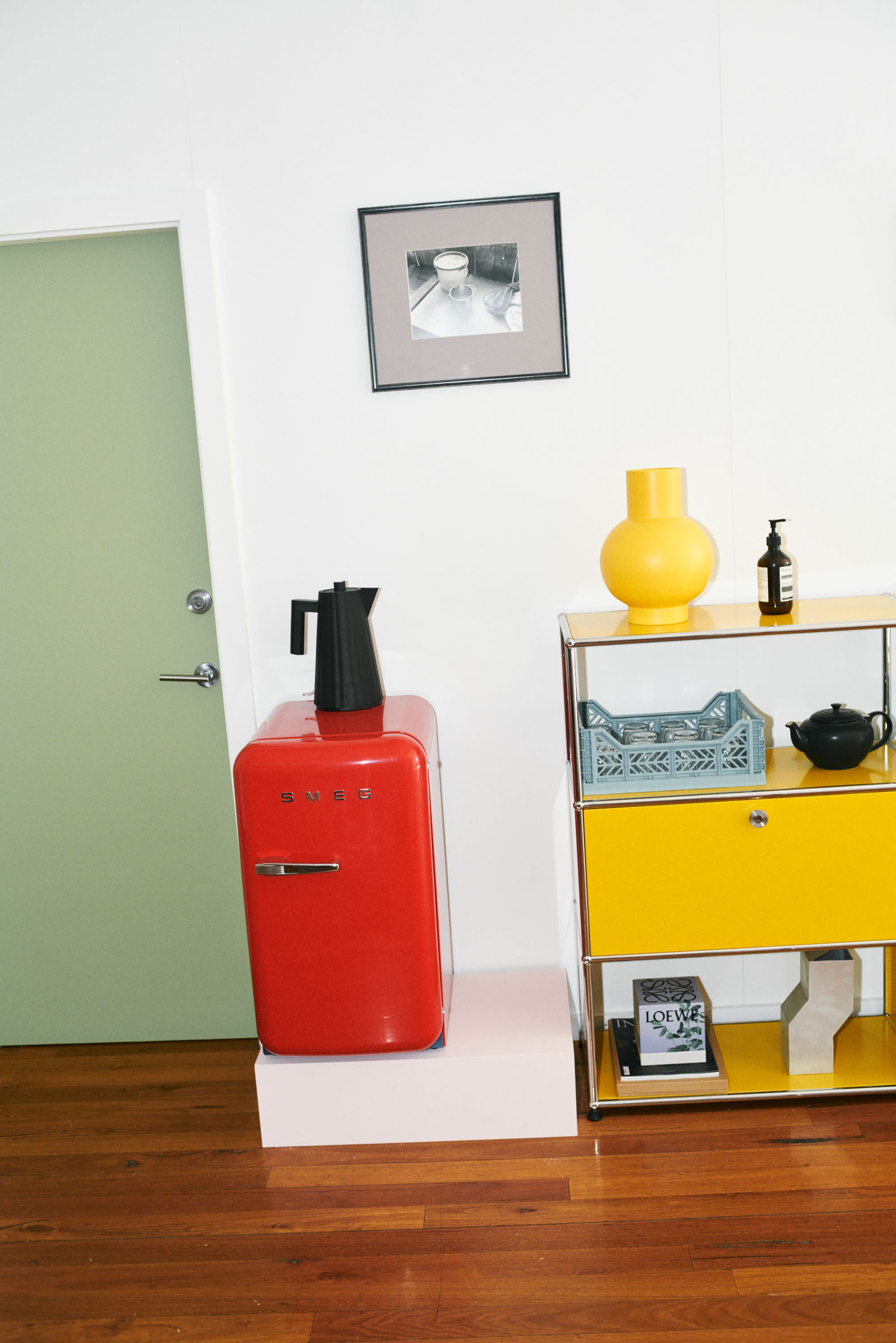 A red retro refrigerator is positioned beside a yellow cabinet against a white wall, with a green door in the background and various decorative items on the cabinet.
