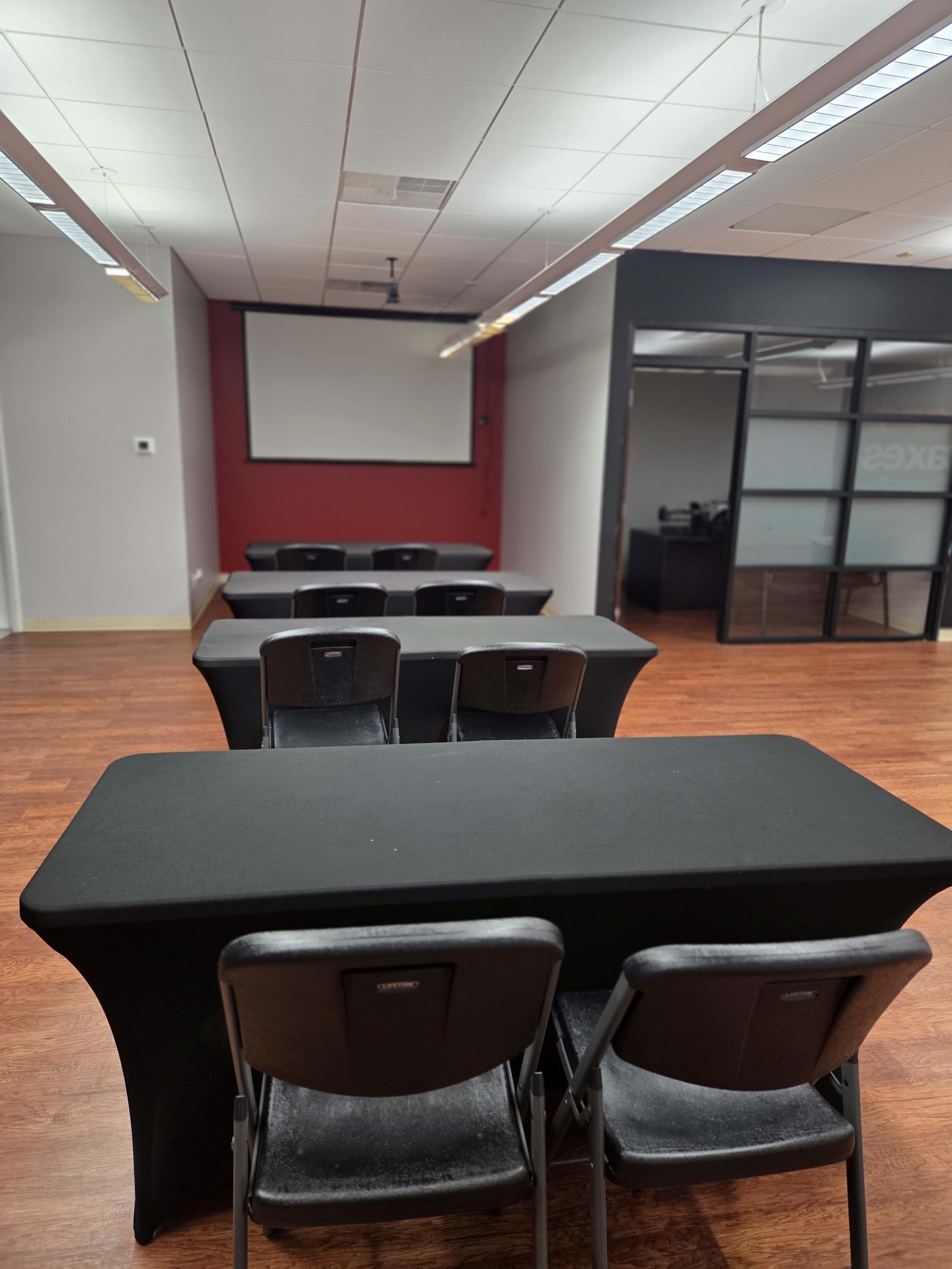 The image shows a meeting room with several rows of black tables and chairs facing a blank wall with a projector screen.