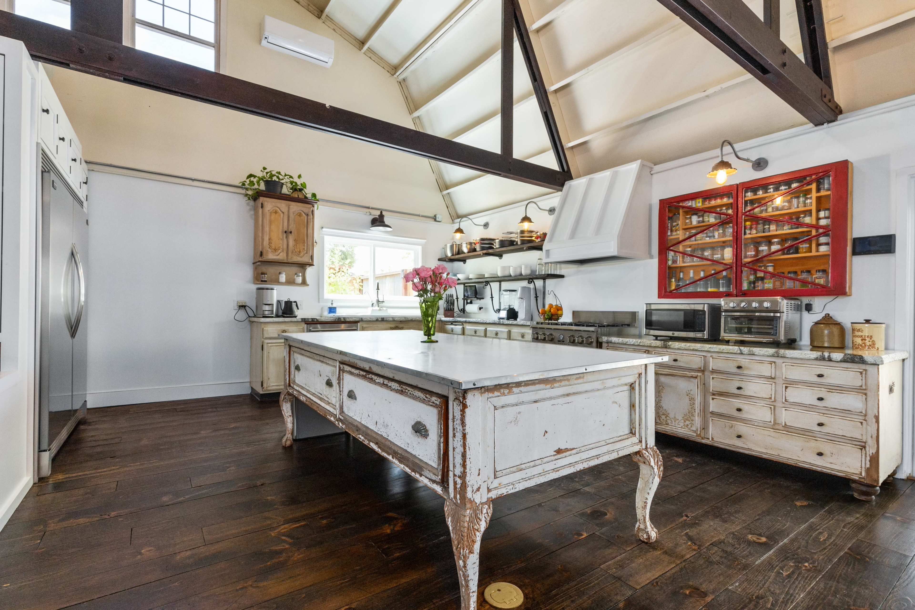 The image shows a spacious kitchen with a large central island, wooden floors, and high, slanted ceilings, featuring a mix of modern and vintage appliances.