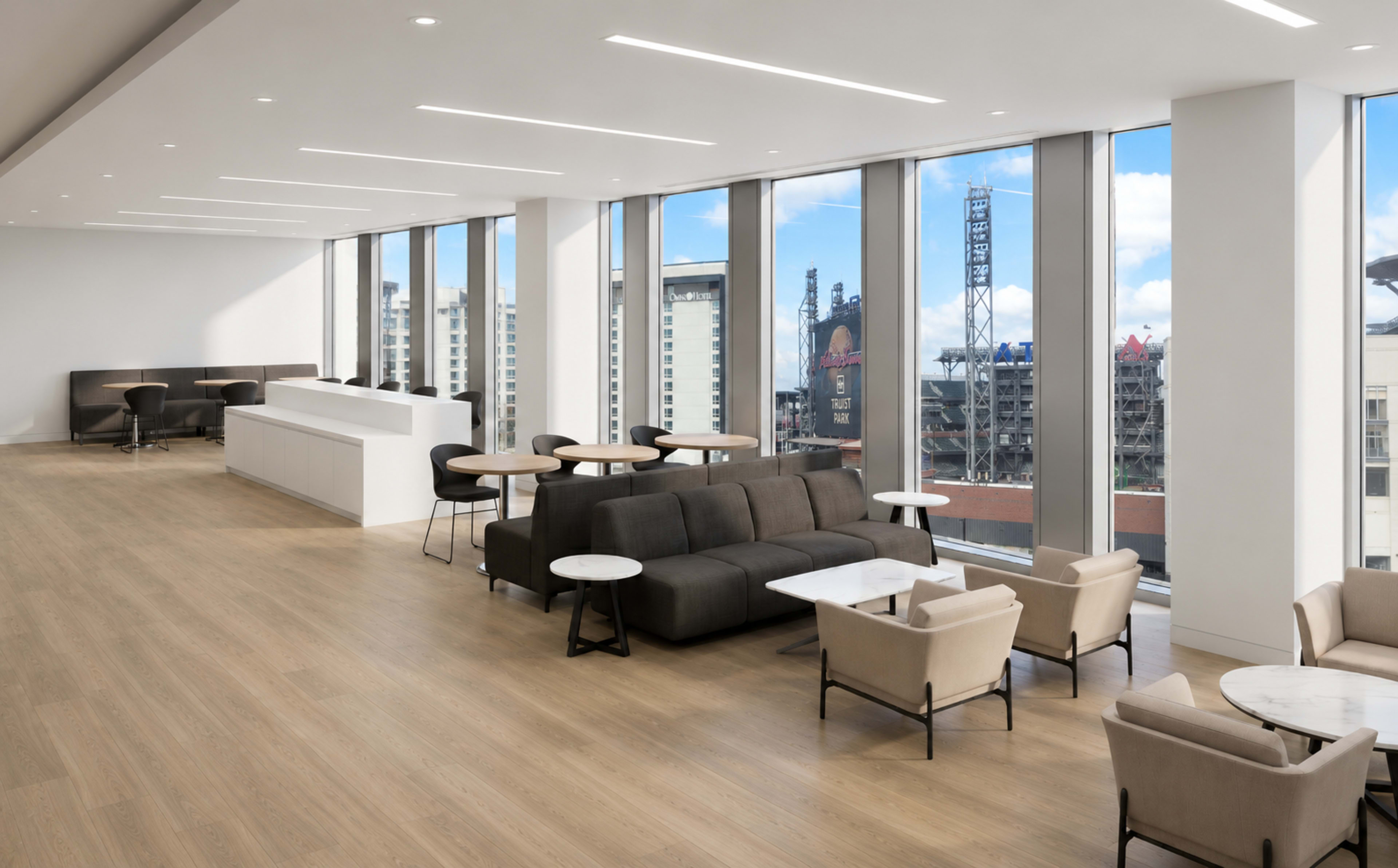 The image shows a modern, spacious lounge area with large windows, showcasing a view of a city skyline, and furnished with various seating arrangements, including sofas and tables.