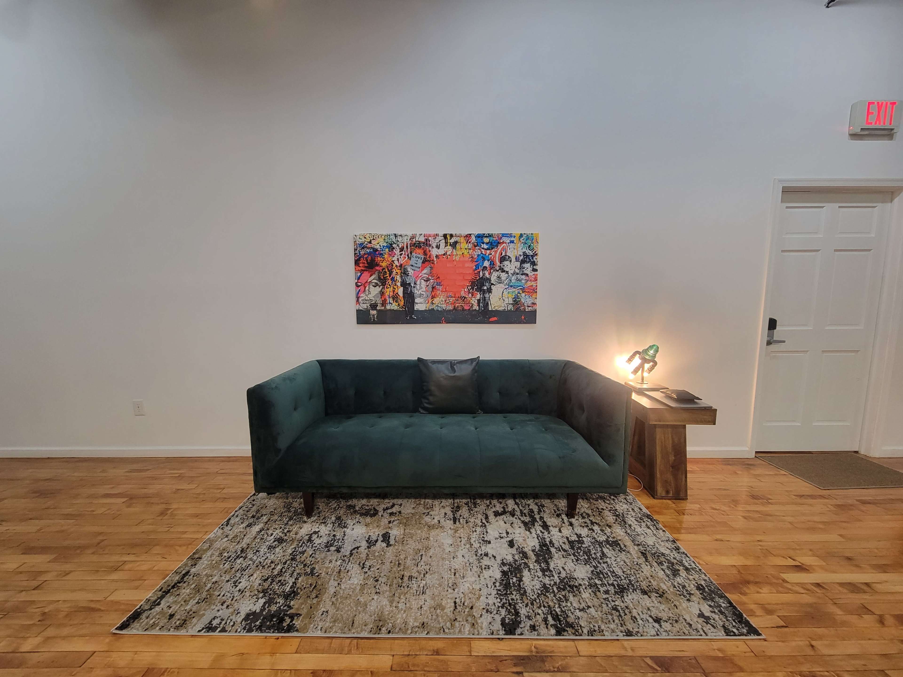 The image shows a green velvet sofa positioned in front of a colorful abstract painting, with a wooden side table and a lamp nearby, all situated on a patterned rug within a spacious, minimalist room.