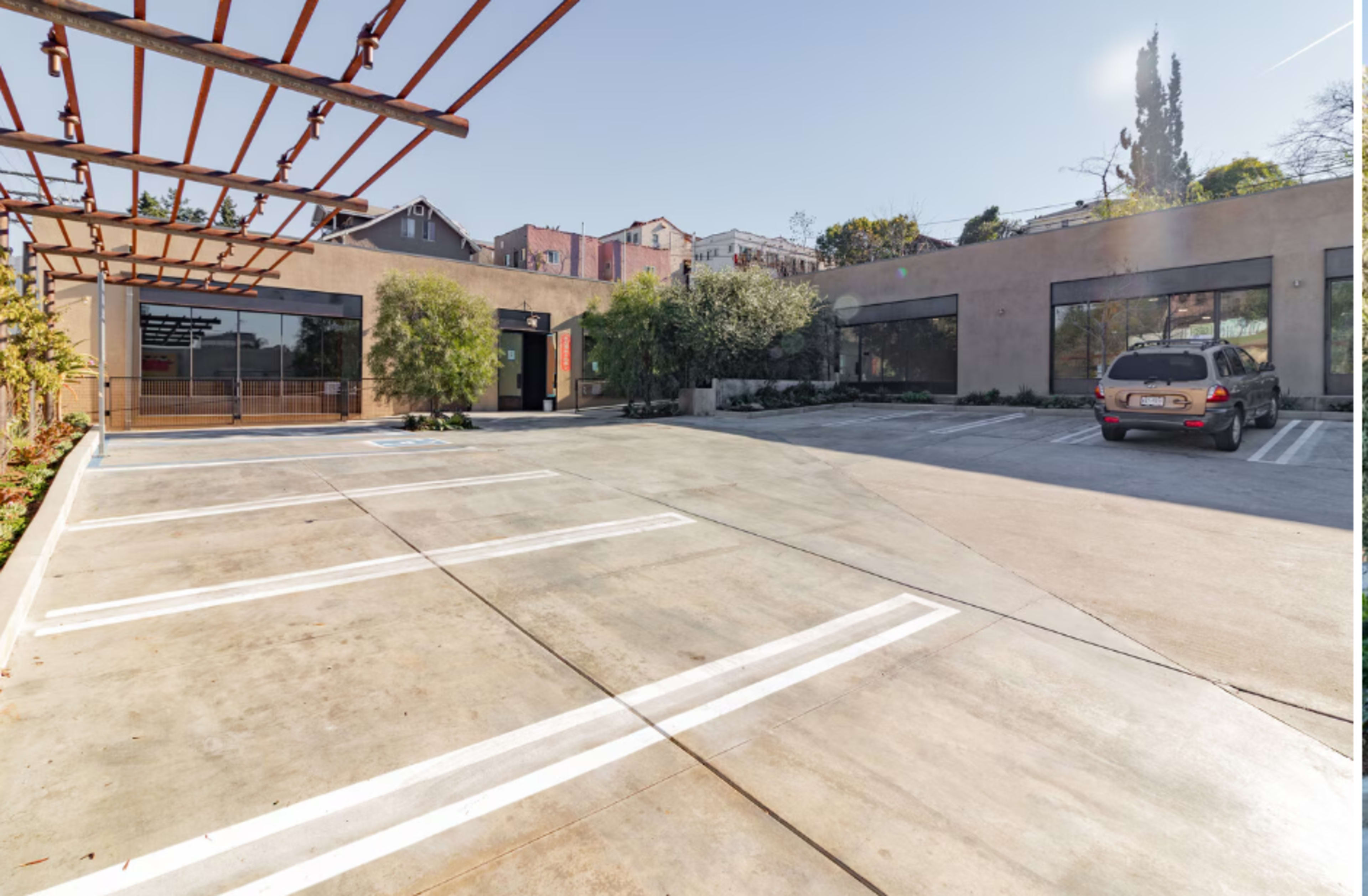 The image shows a commercial building with large glass windows and a paved parking lot, featuring a car and a trellis structure on one side.