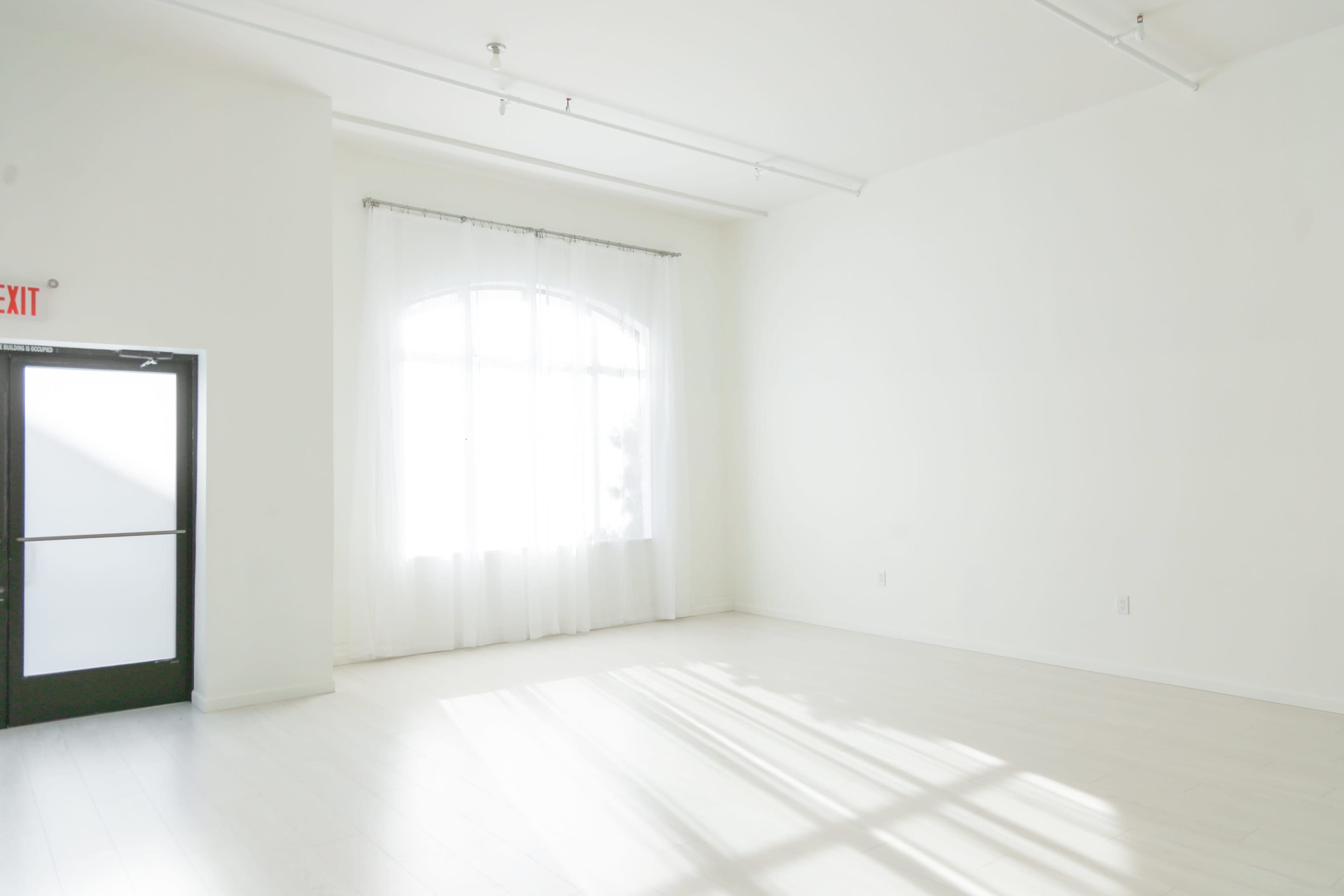The image shows a minimally furnished, bright room with large windows, white walls, and light-colored flooring.
