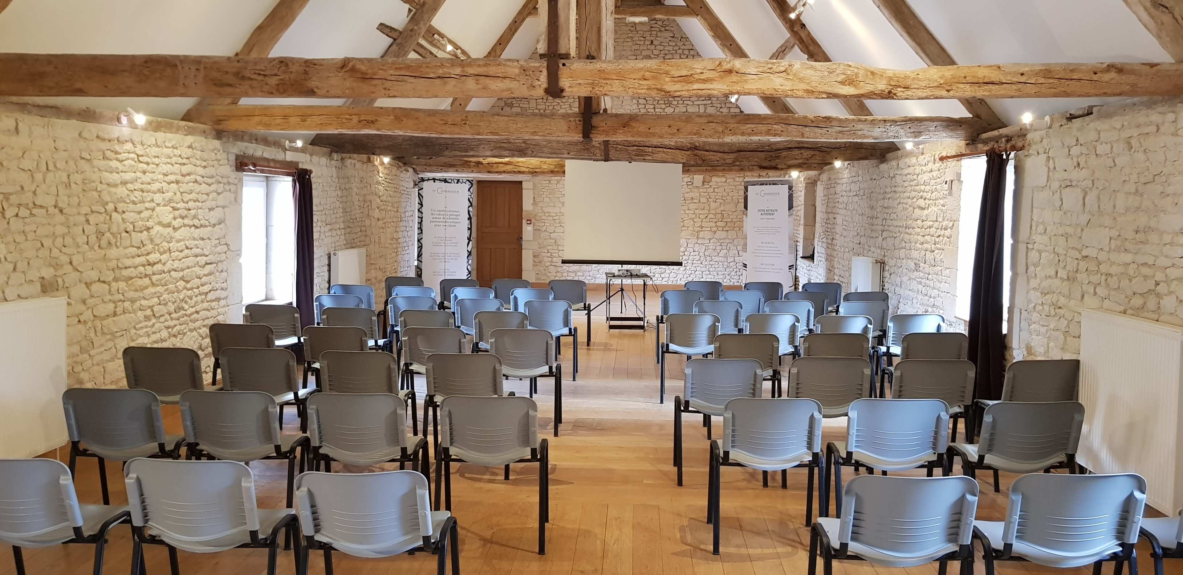 An empty meeting room features rows of blue chairs facing a projector screen, with exposed wooden beams and stone walls.
