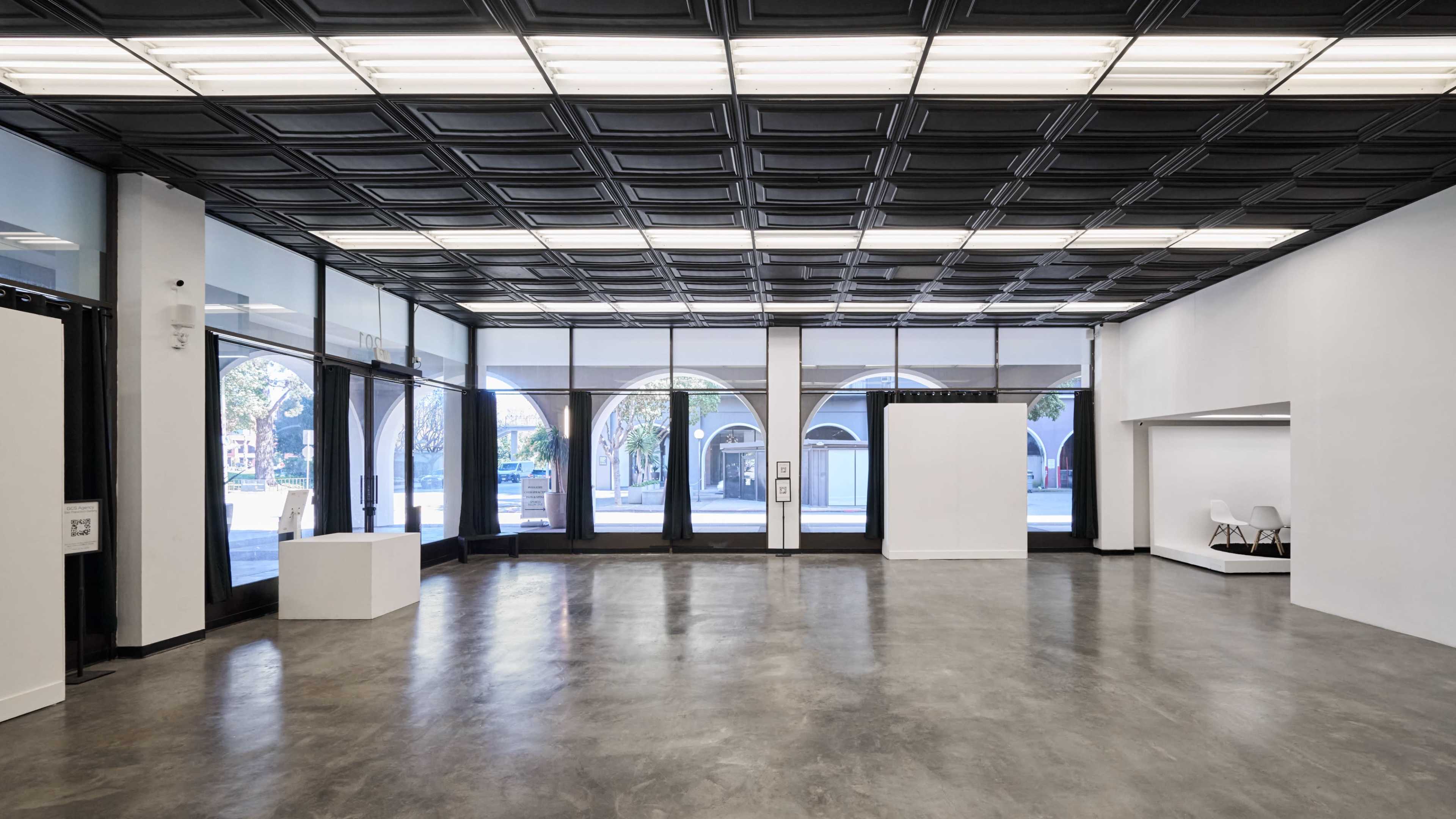 The image shows a spacious, well-lit interior of an art gallery with large windows and a black ceiling.