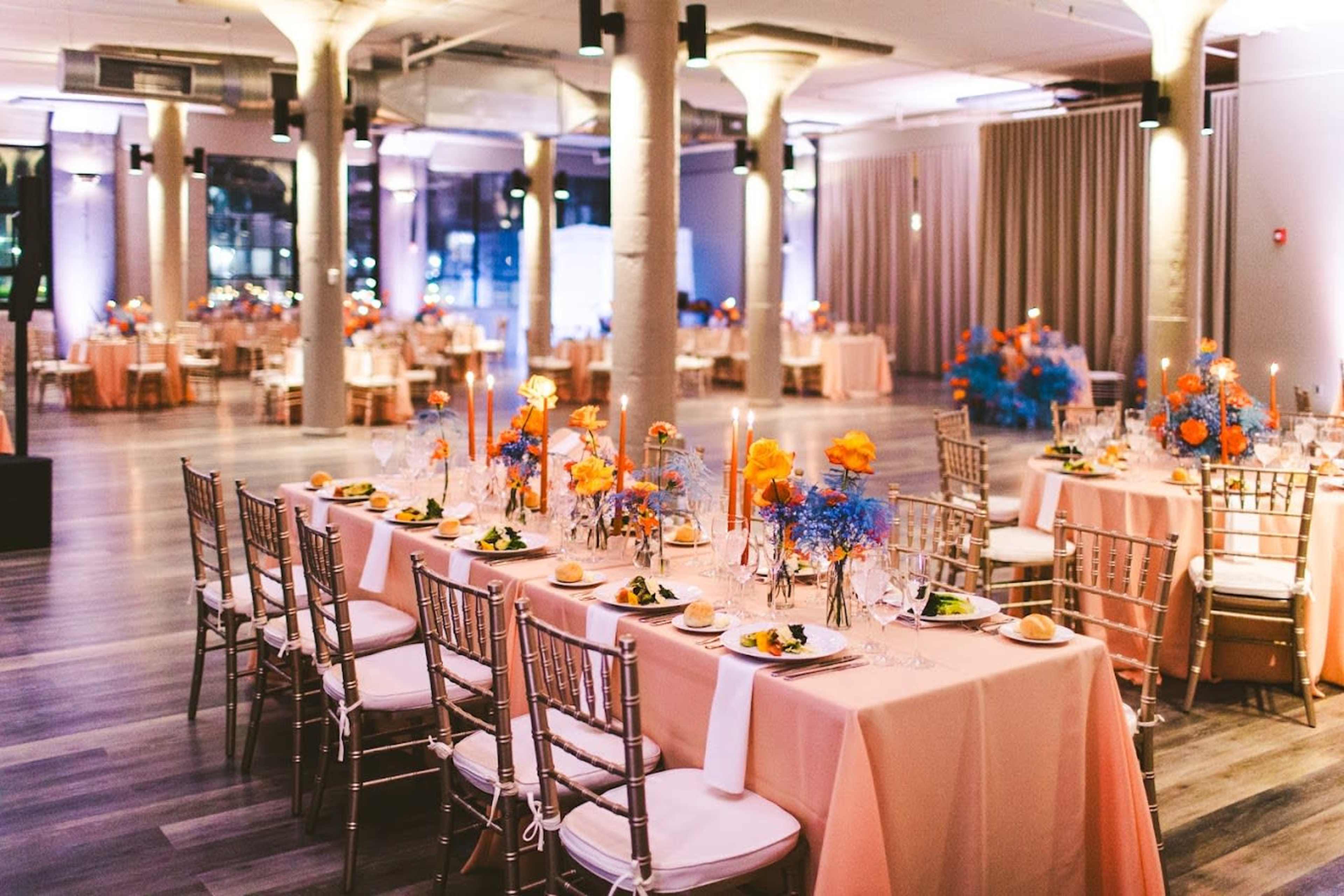 A spacious event hall is set up with elegantly arranged tables adorned with pink tablecloths and vibrant floral centerpieces.
