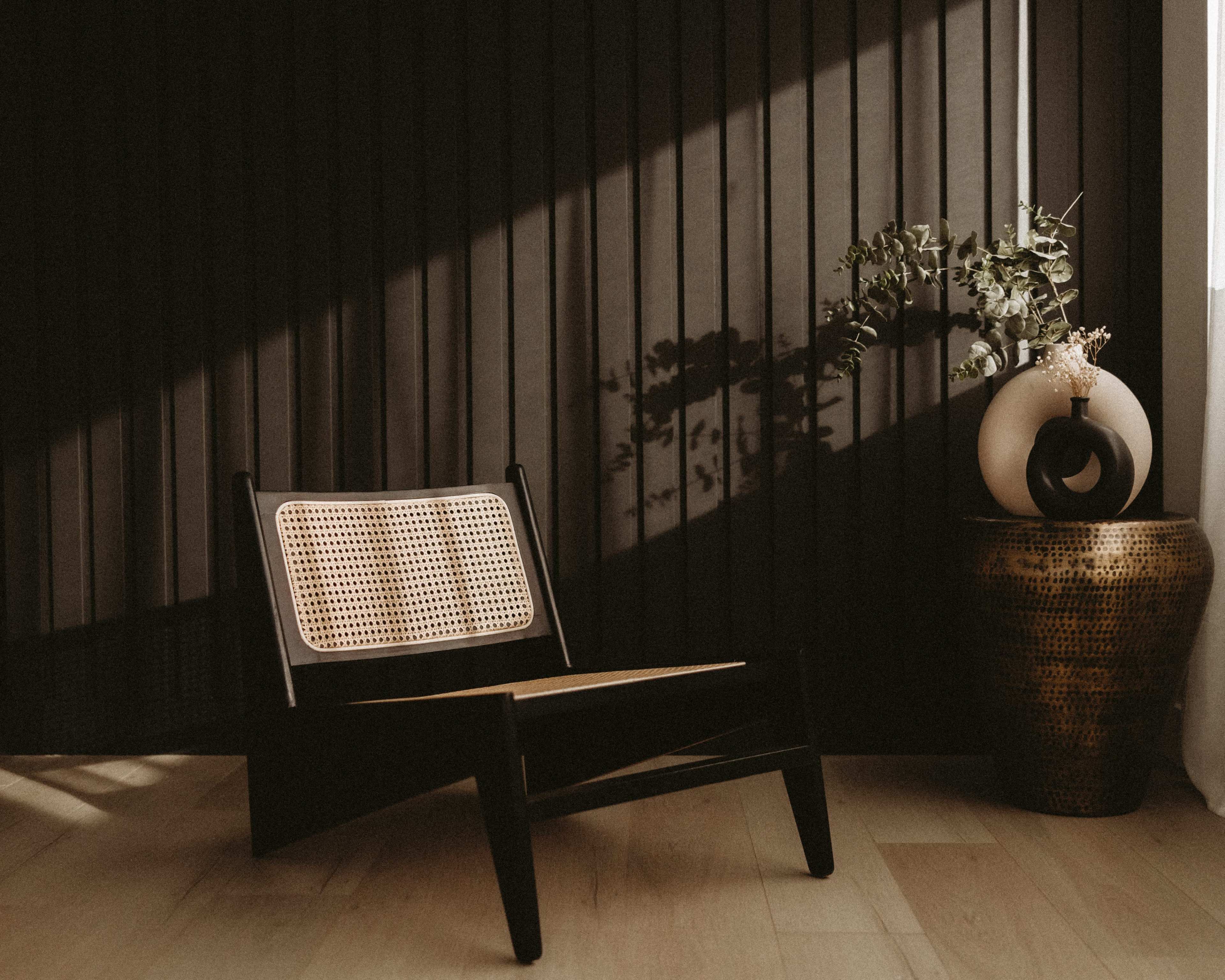 A minimalist room features a black chair with a woven backrest next to a decorative vase and a potted plant against a slatted wall.