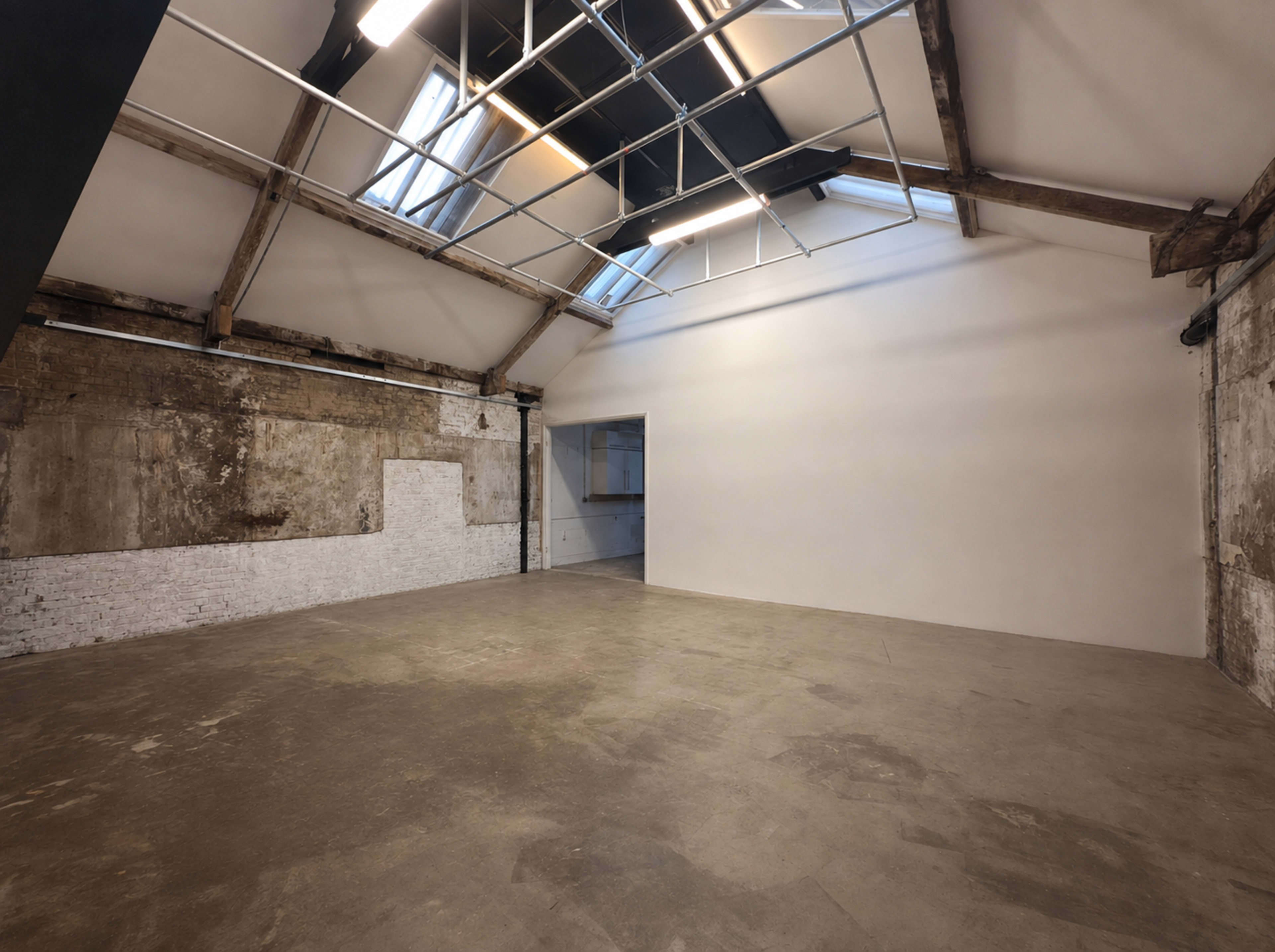 The image shows an open, empty studio space with exposed brick walls, large windows, and a high ceiling featuring wooden beams.