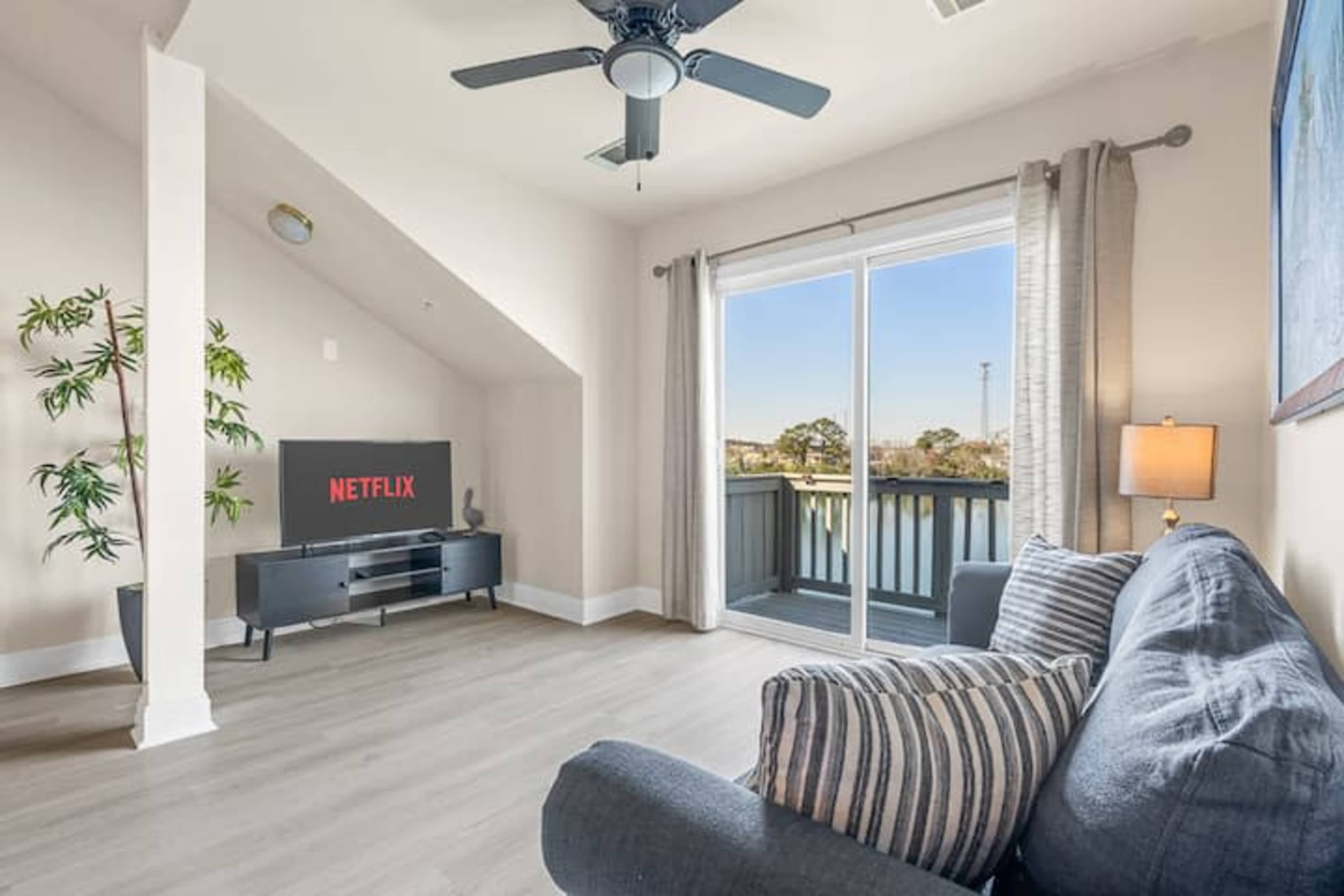 The image shows a cozy living room featuring a dark gray sofa, a television displaying the Netflix logo, and a balcony with large glass doors allowing light to enter the space.