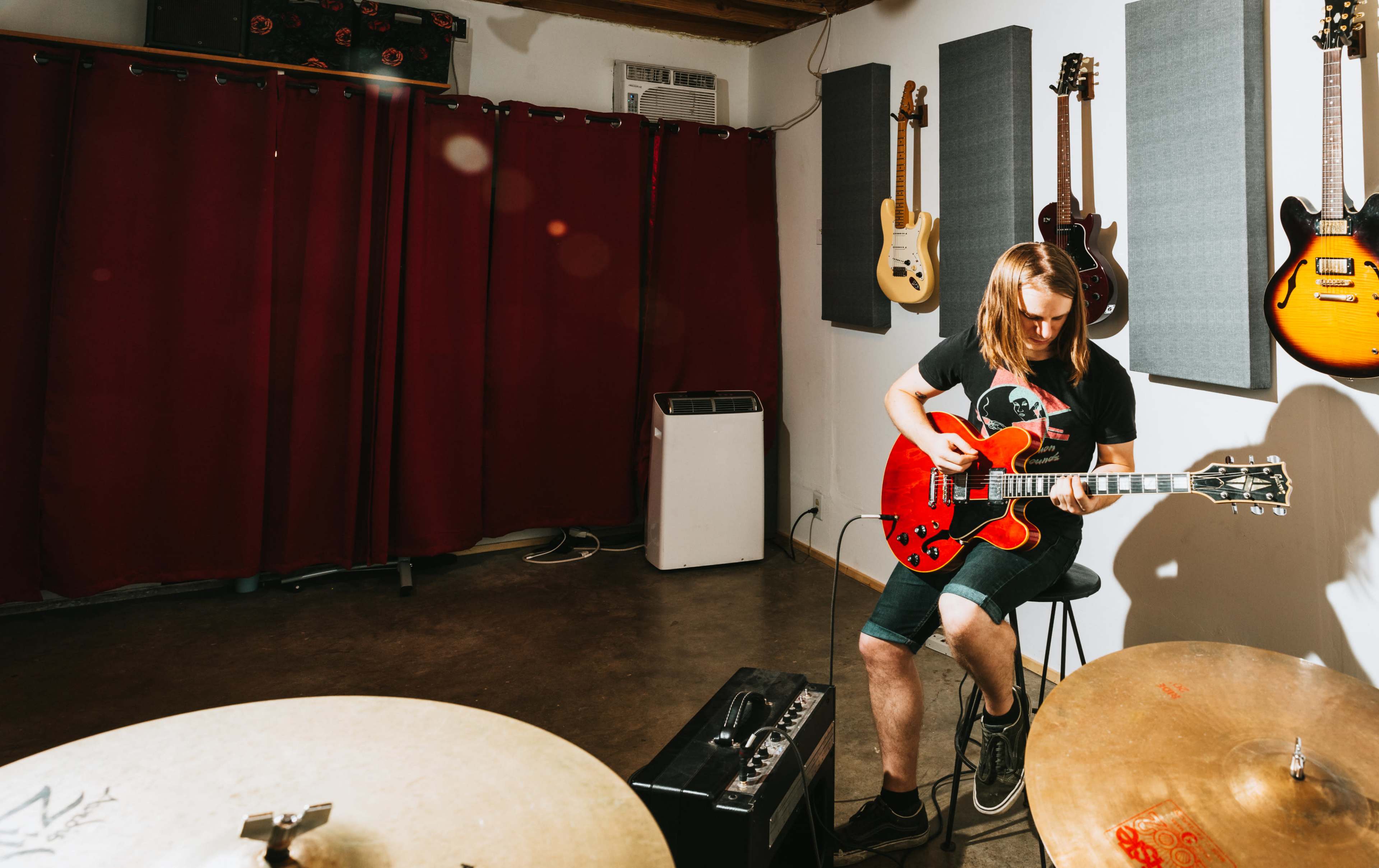 A musician plays an electric guitar while sitting on a stool in a room featuring several hanging guitars and soundproofing panels.