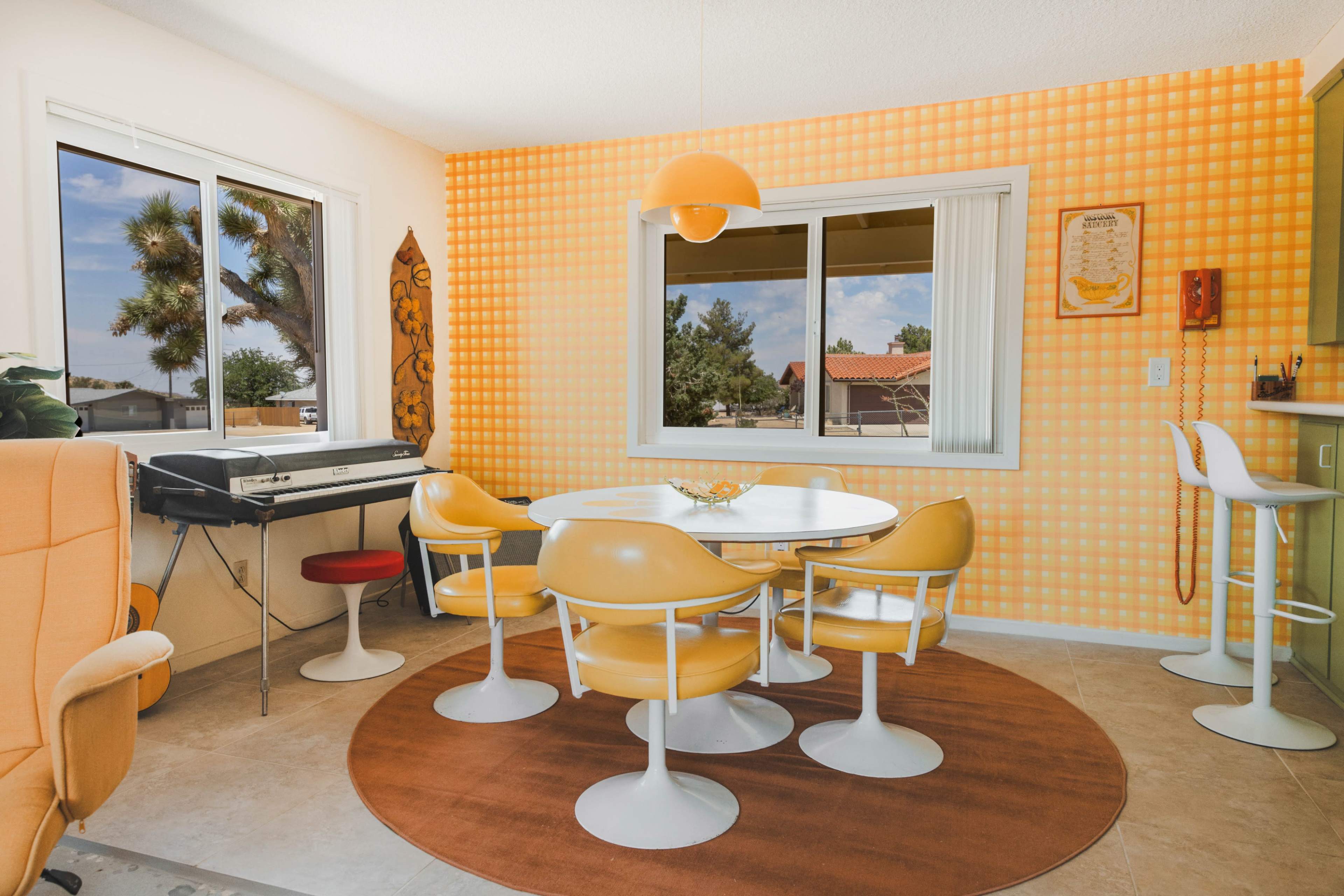 A retro-style dining area features a round table surrounded by yellow chairs, an orange carpet, and an orange wall with a large window.