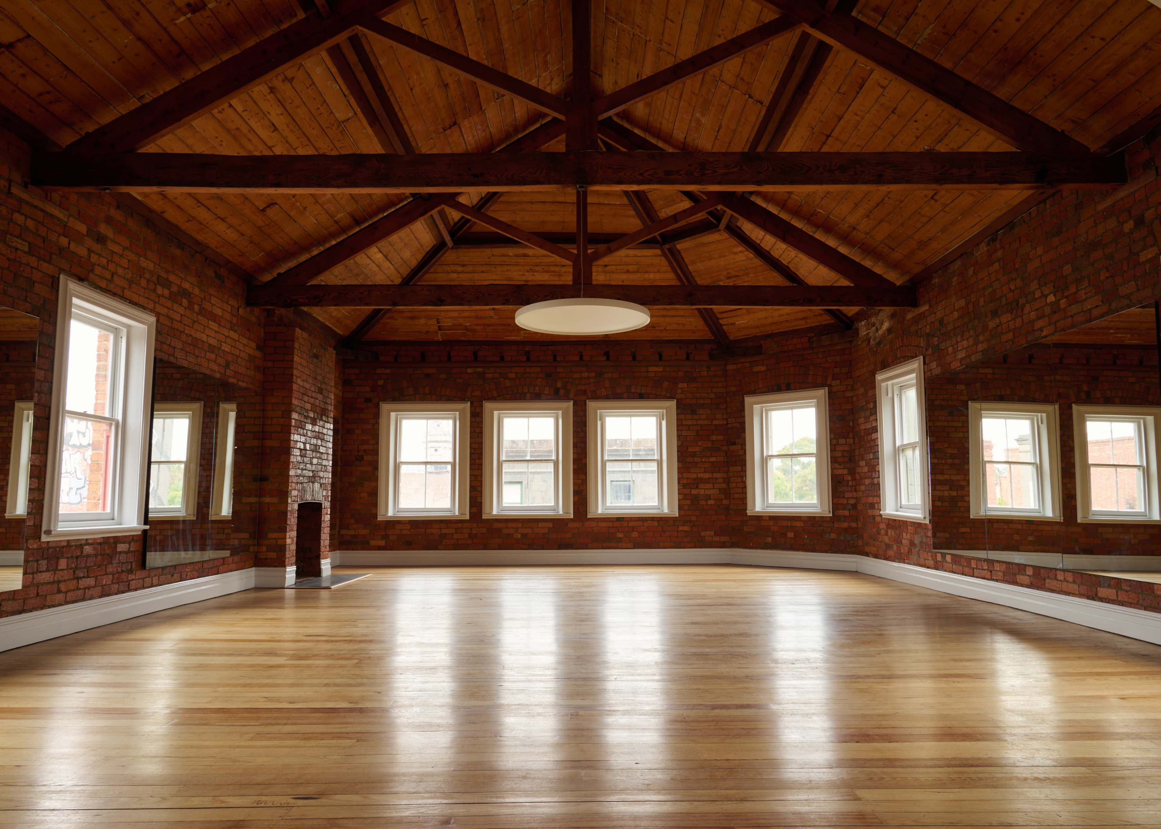 The image shows a spacious room with wooden beams, brick walls, large windows, and a polished wooden floor.