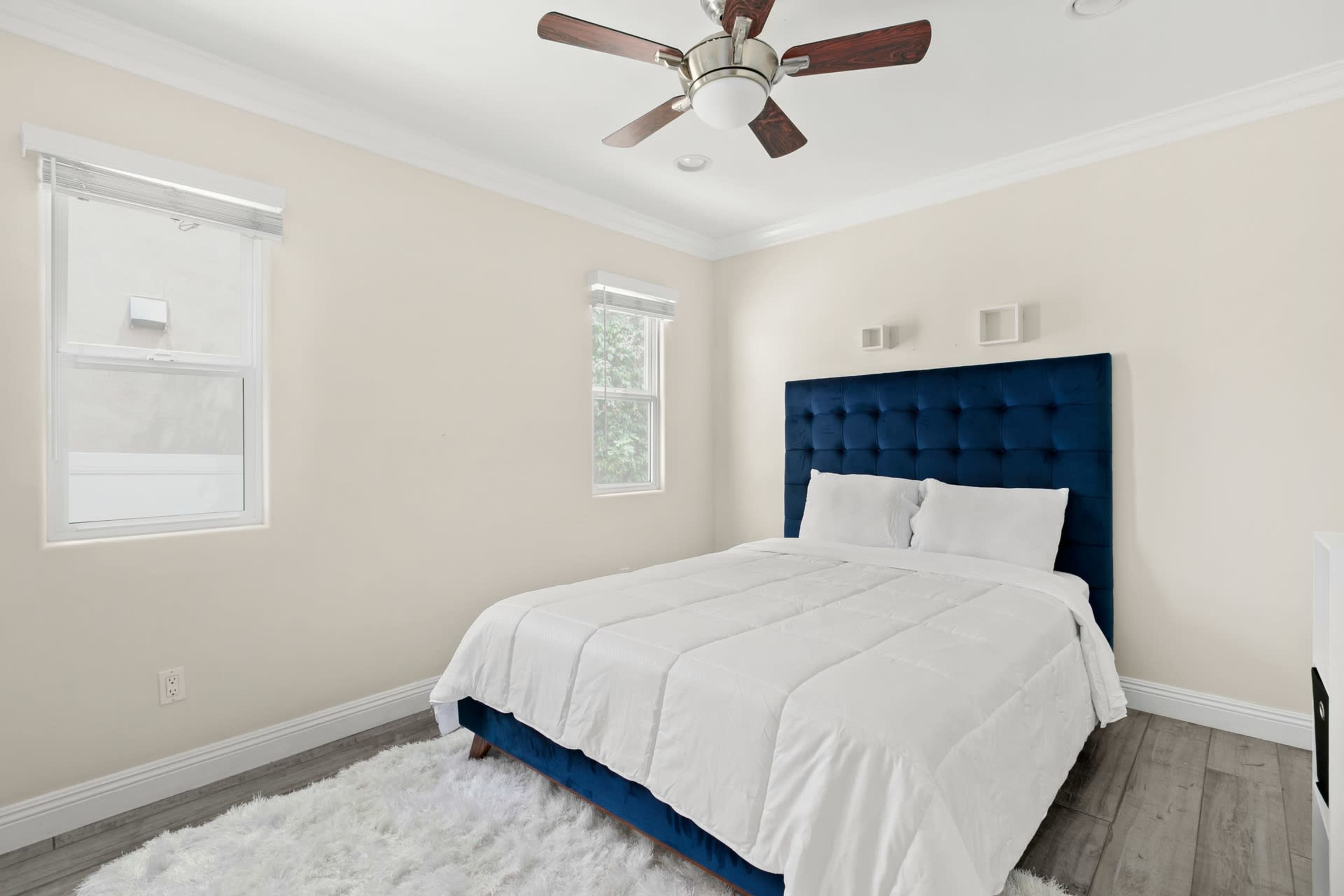 The image shows a neatly arranged bedroom featuring a blue tufted headboard, a white bedspread, and two windows with blinds.