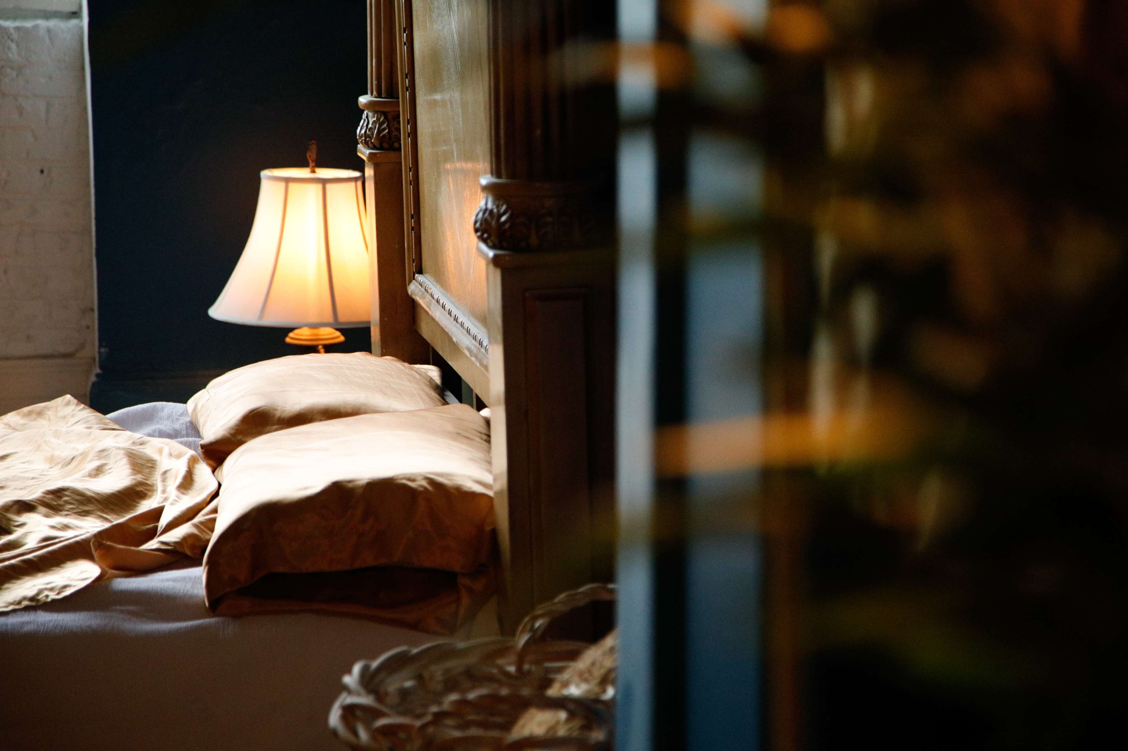 A softly lit bedroom features a bed with two beige pillows, partially covered by a light-colored blanket, and a bedside lamp casting a warm glow.