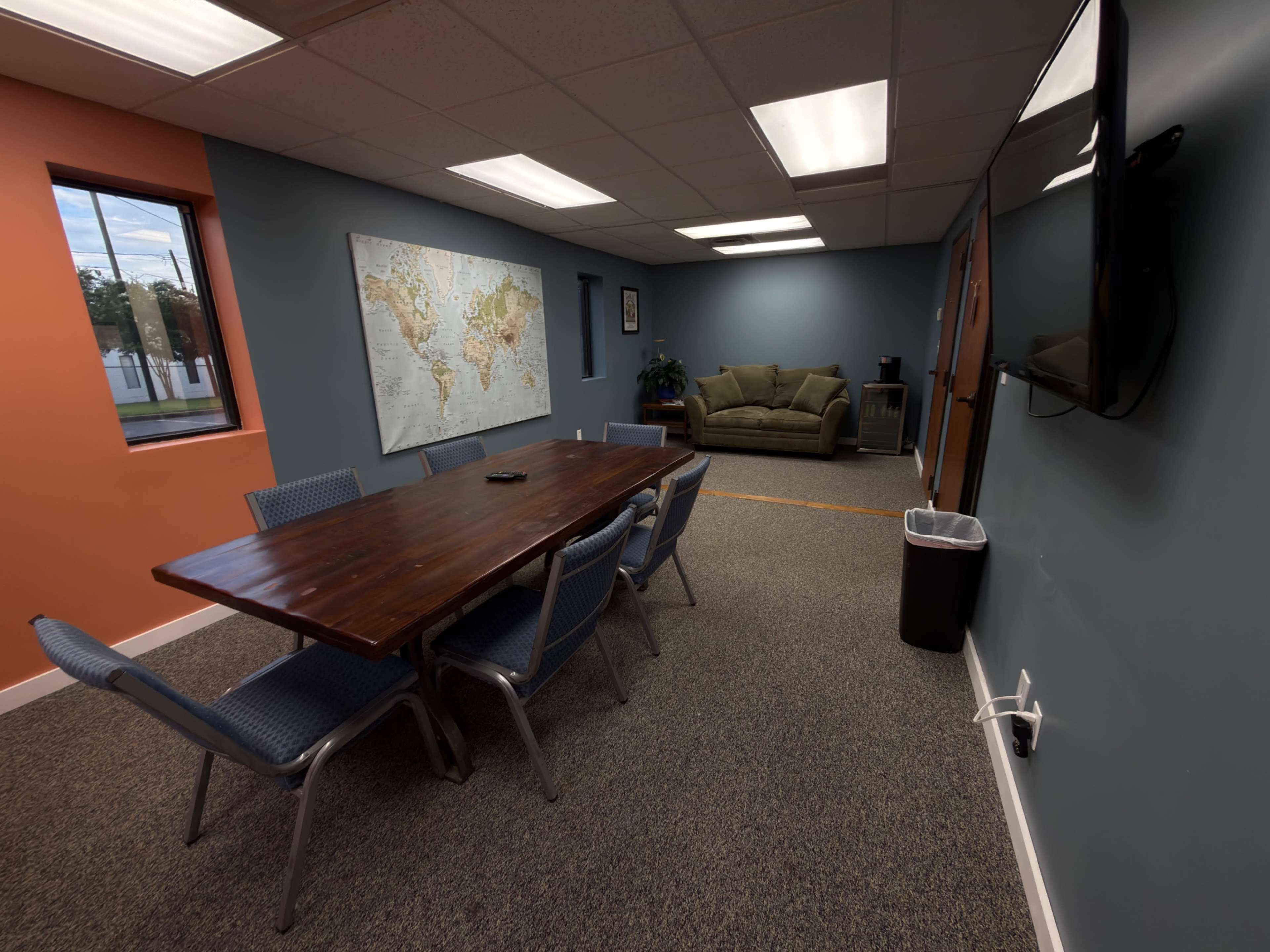 The image shows a meeting room with a large wooden table surrounded by chairs, a world map on the wall, and a couch in the corner.