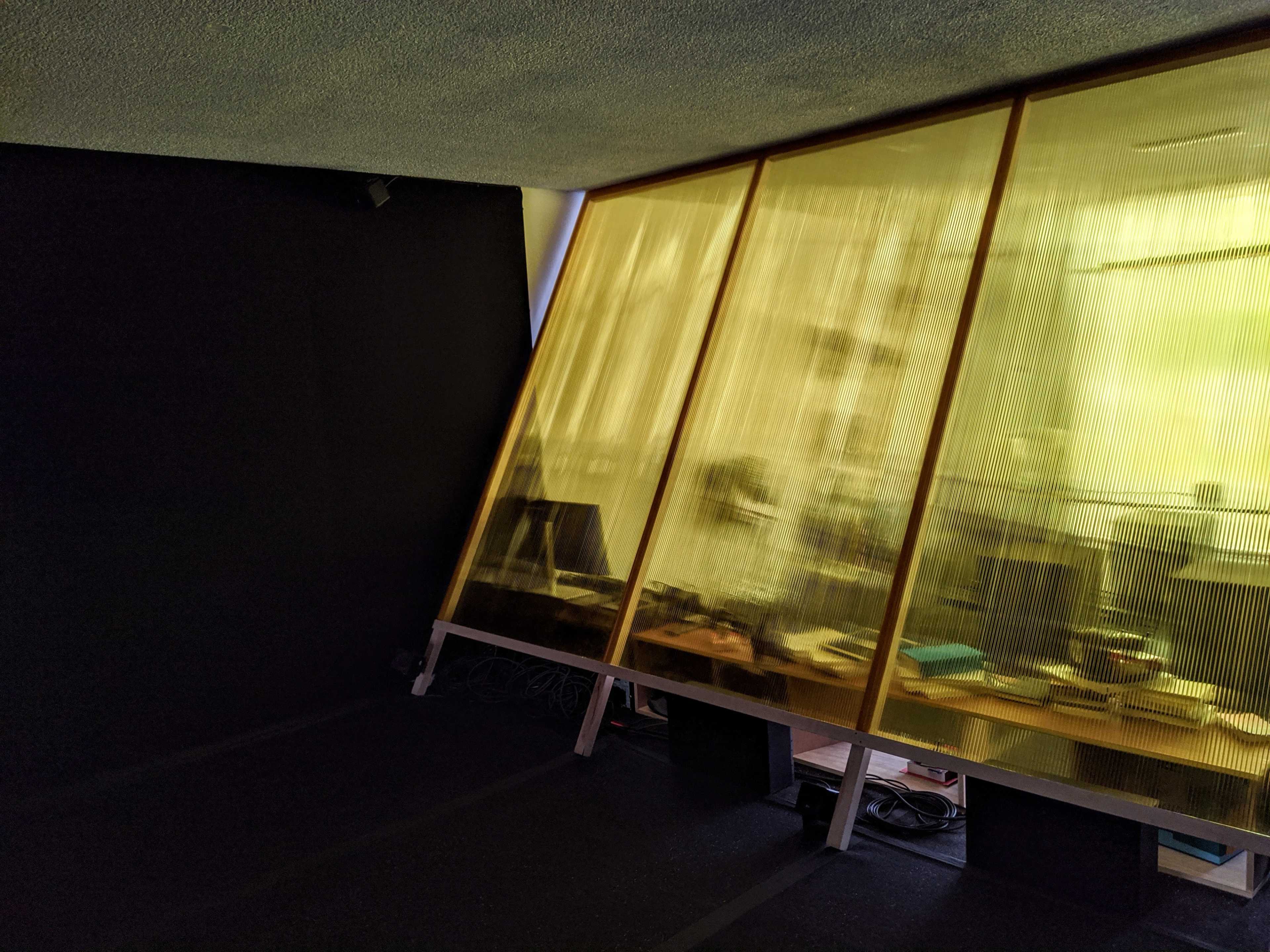 A room features a large, angled window with yellow-tinted panels, revealing a workspace filled with desks and equipment.