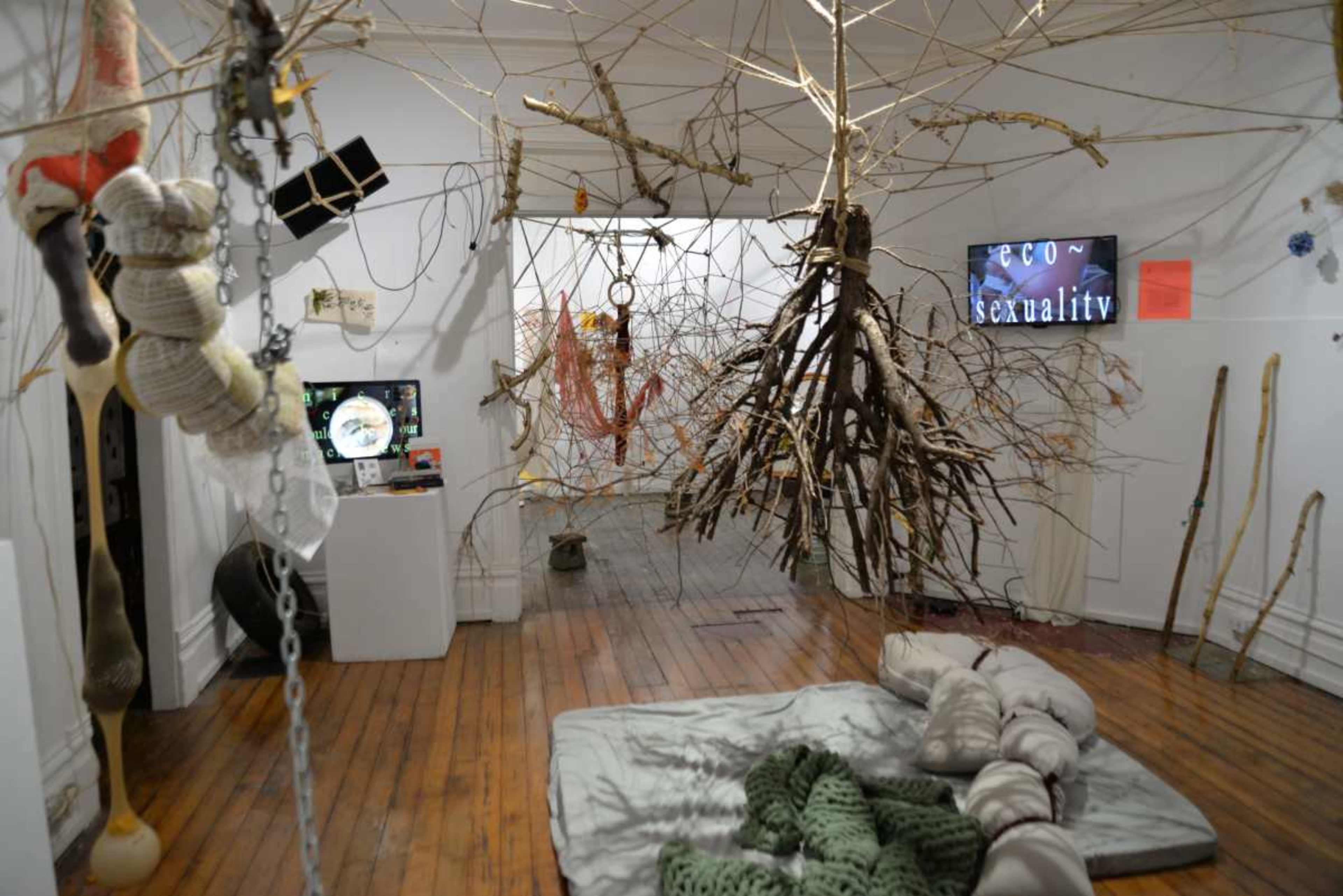An art installation featuring various branches and twigs suspended from the ceiling, with a mixture of textiles on the floor and a screen displaying text in the background.