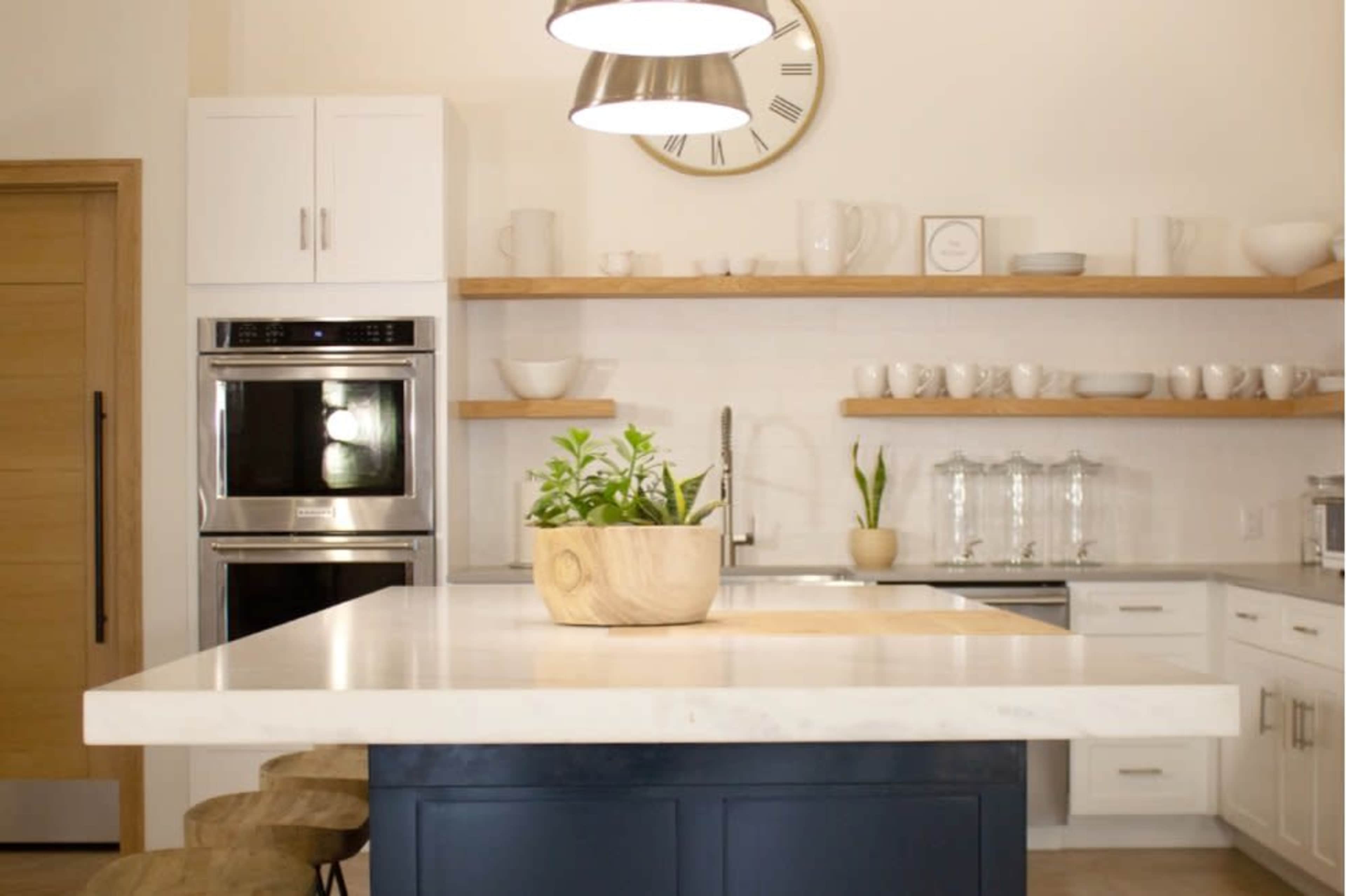 A modern kitchen features a central island with a plant in a wooden bowl, stainless steel appliances, and open shelves displaying dishware.