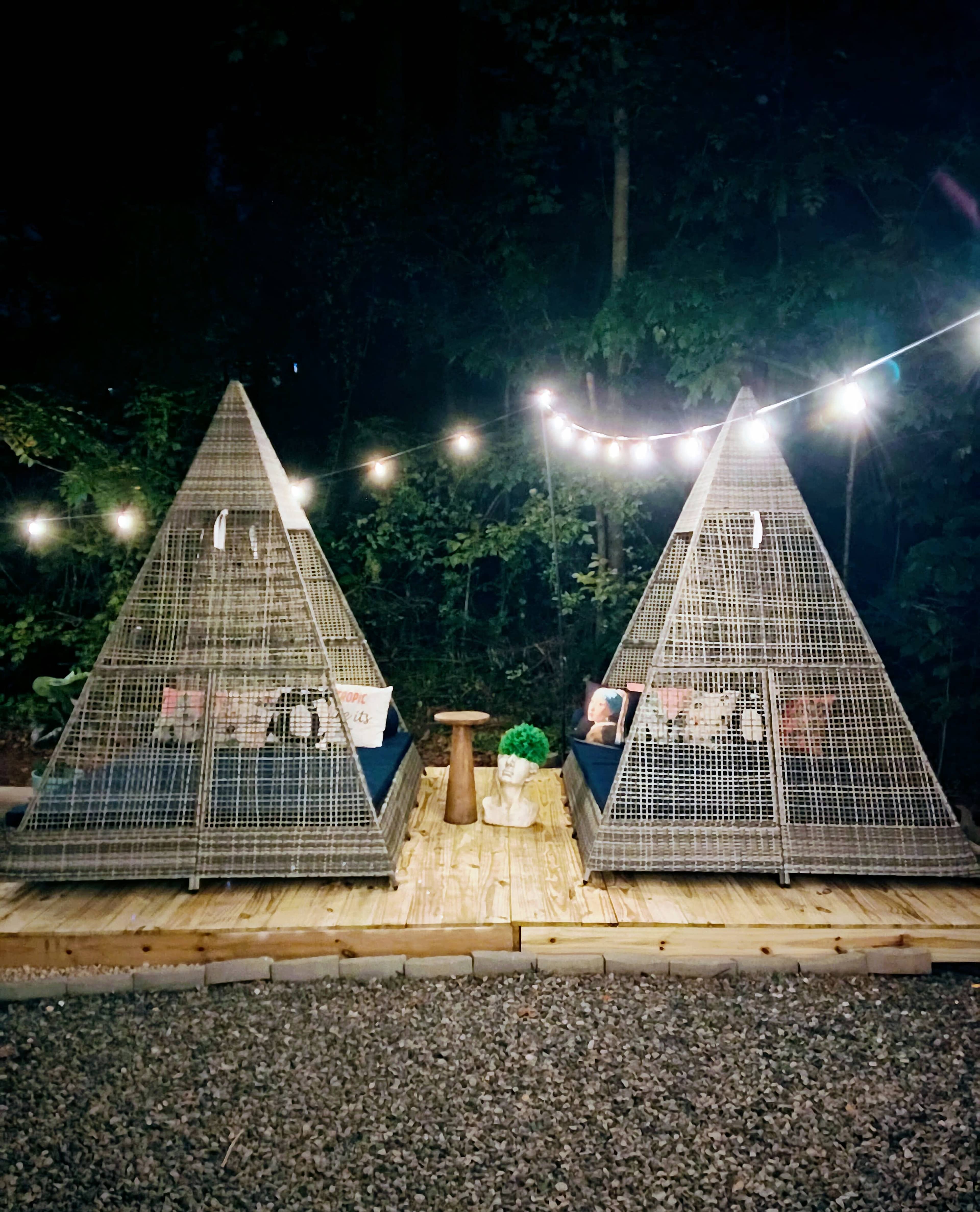 The image shows two triangular-shaped wire structures placed on a wooden deck, illuminated by string lights in a wooded area at night.