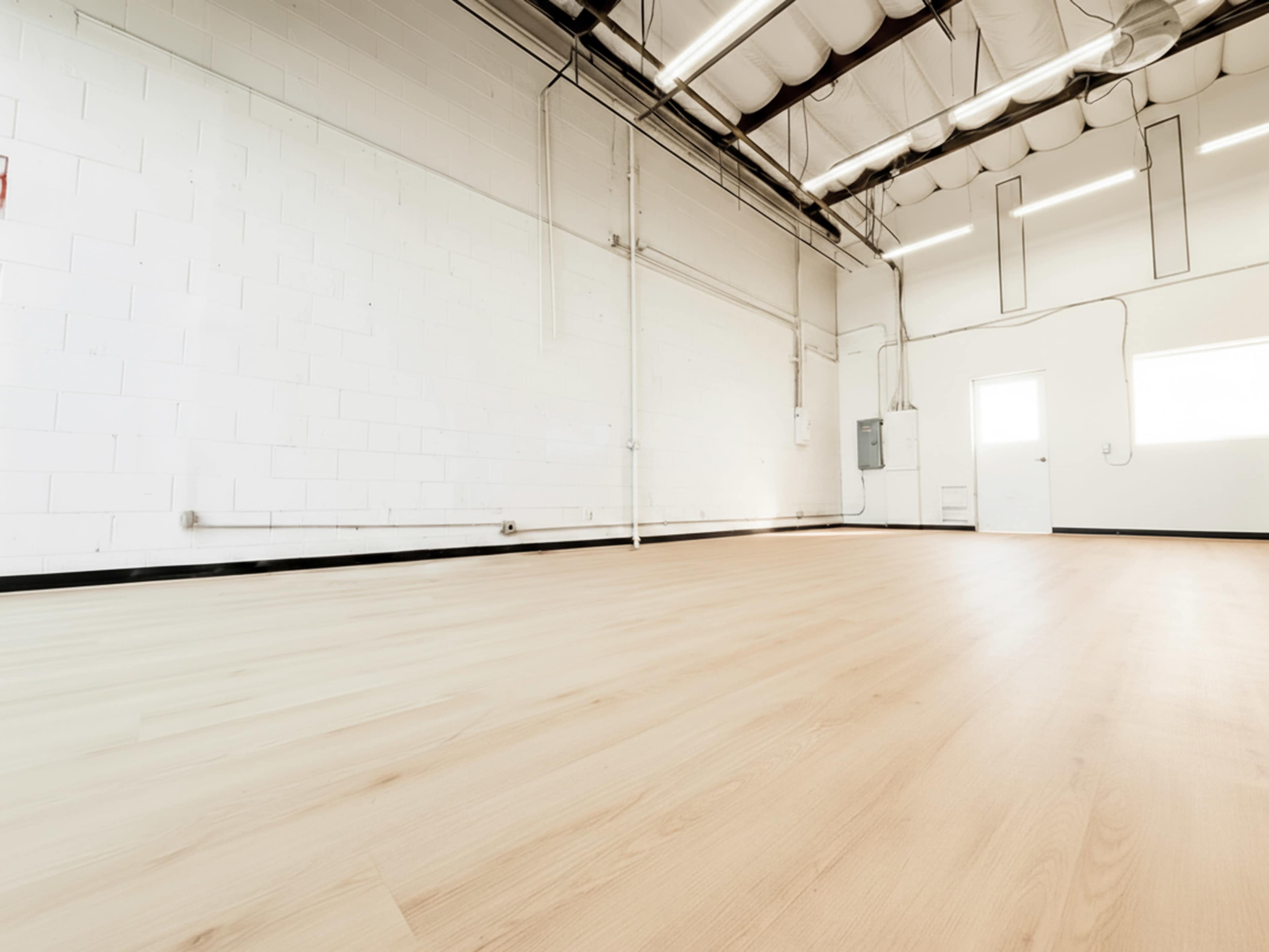 The image shows a spacious, empty room with a wooden floor and white walls, featuring overhead lights and a door at one end.