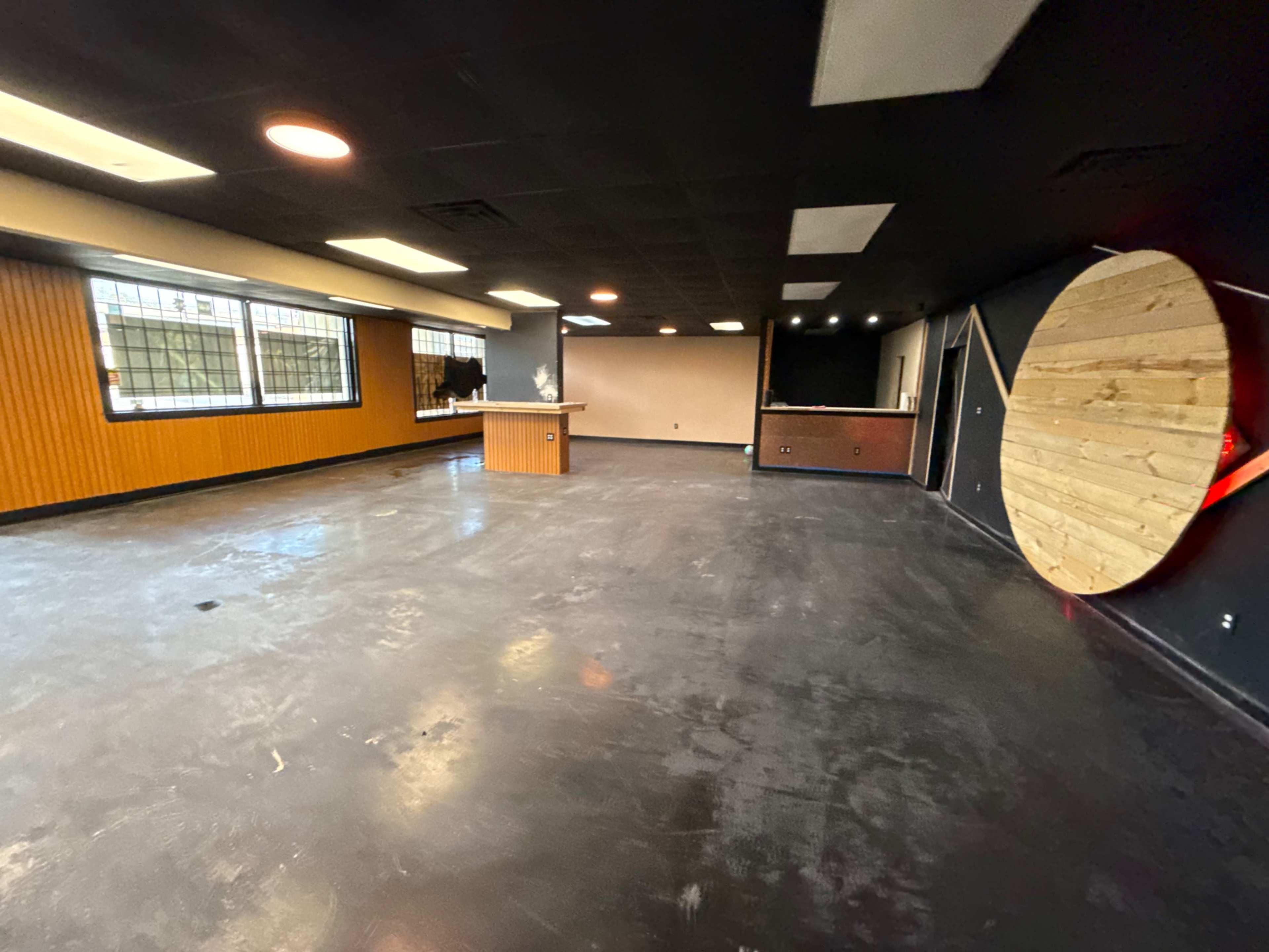 The image shows an empty interior space with a polished concrete floor, a bar area on one side, and a large circular wooden feature on another wall.