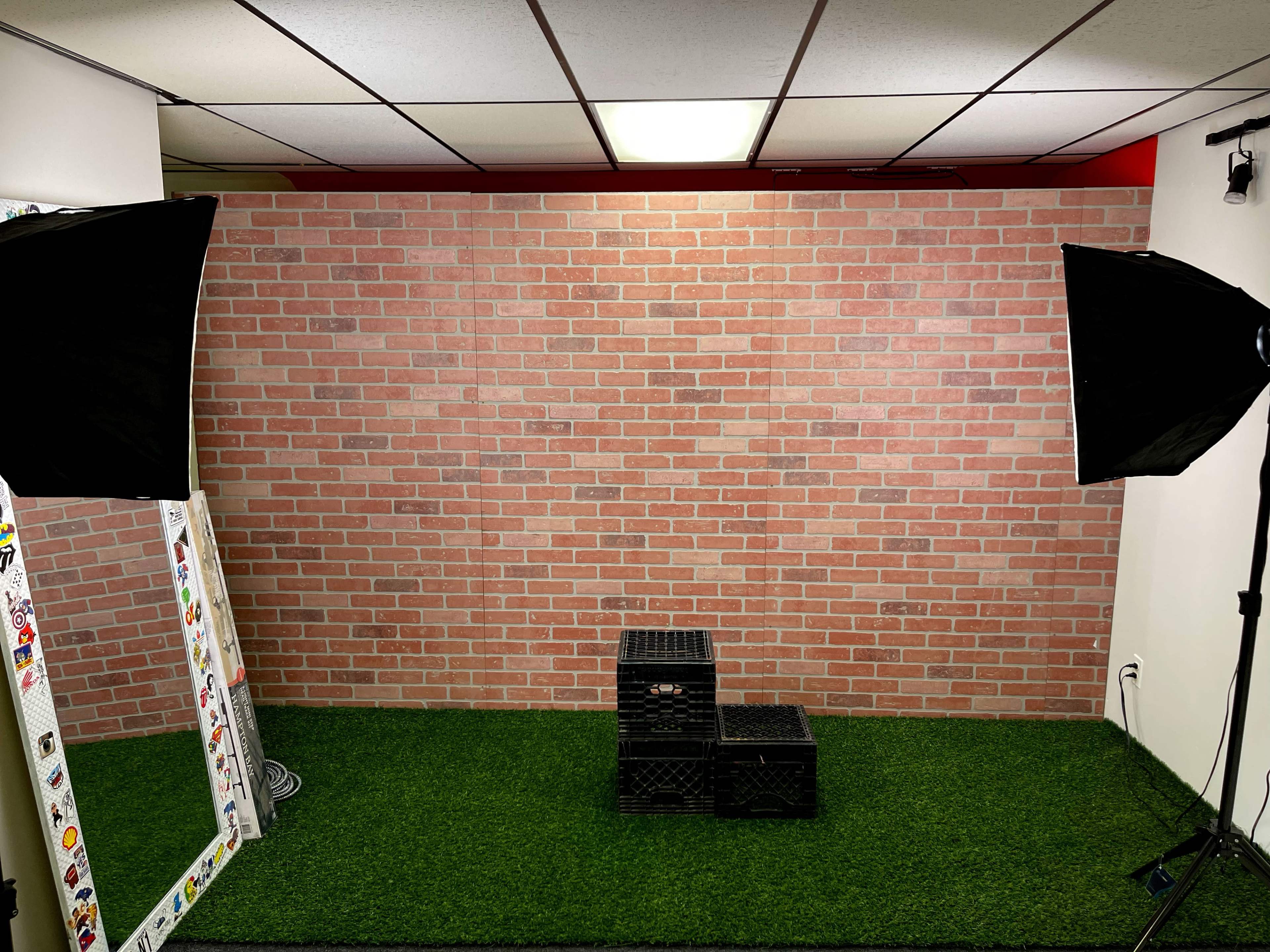 A studio space featuring a brick wall backdrop, two lighting setups, a mirror, and black crates on a green turf floor.