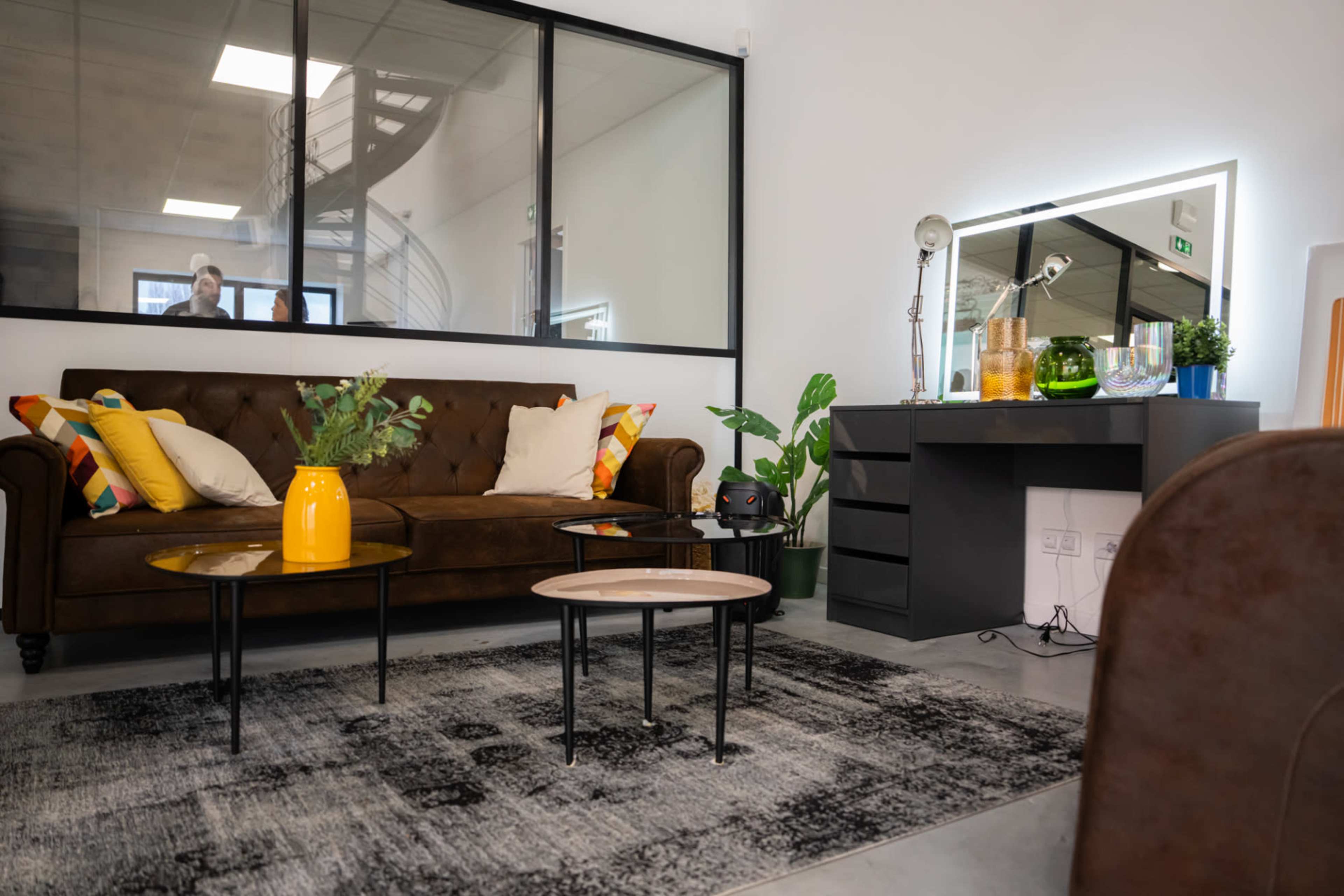 A modern indoor living space featuring a brown sofa with colorful cushions, a coffee table, and a black dresser, set against a backdrop of a glass wall and a staircase.