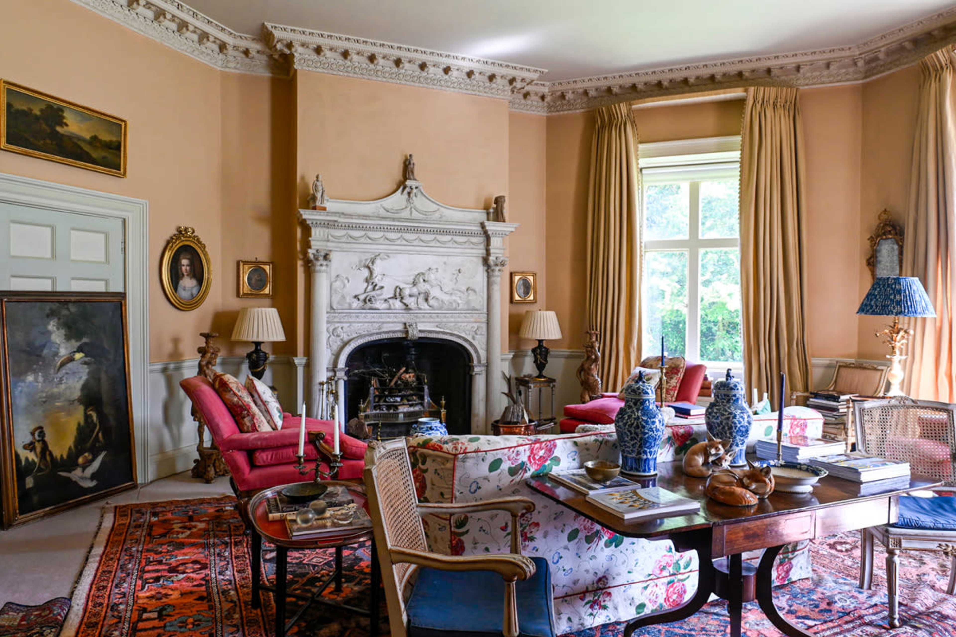 A Victorian-style living room features a stone fireplace, colorful armchairs, and a large window complemented by ornate decorations.