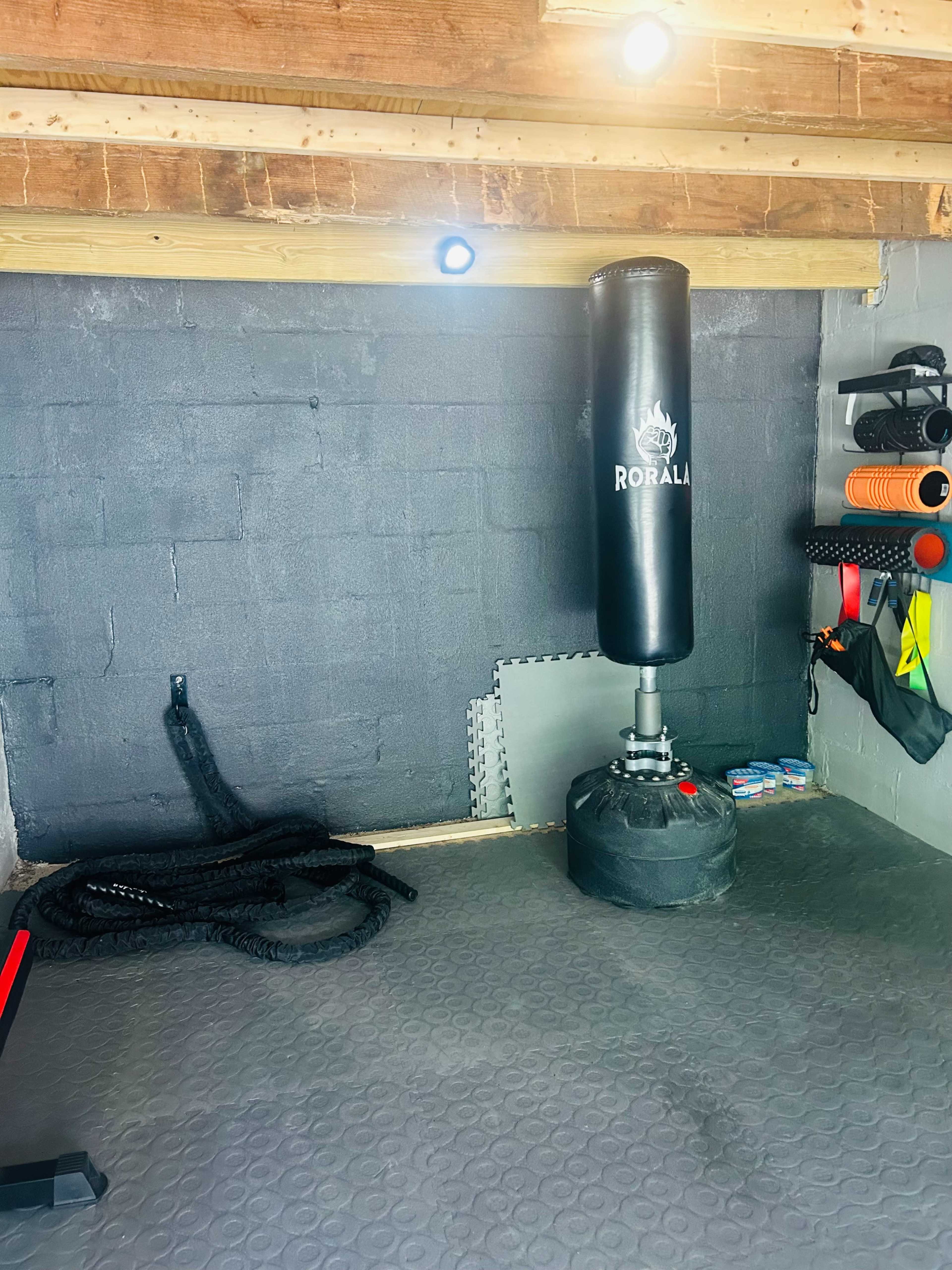 A home gym with a heavy bag suspended from the ceiling, exercise ropes coiled on the floor, and various fitness equipment attached to the wall.