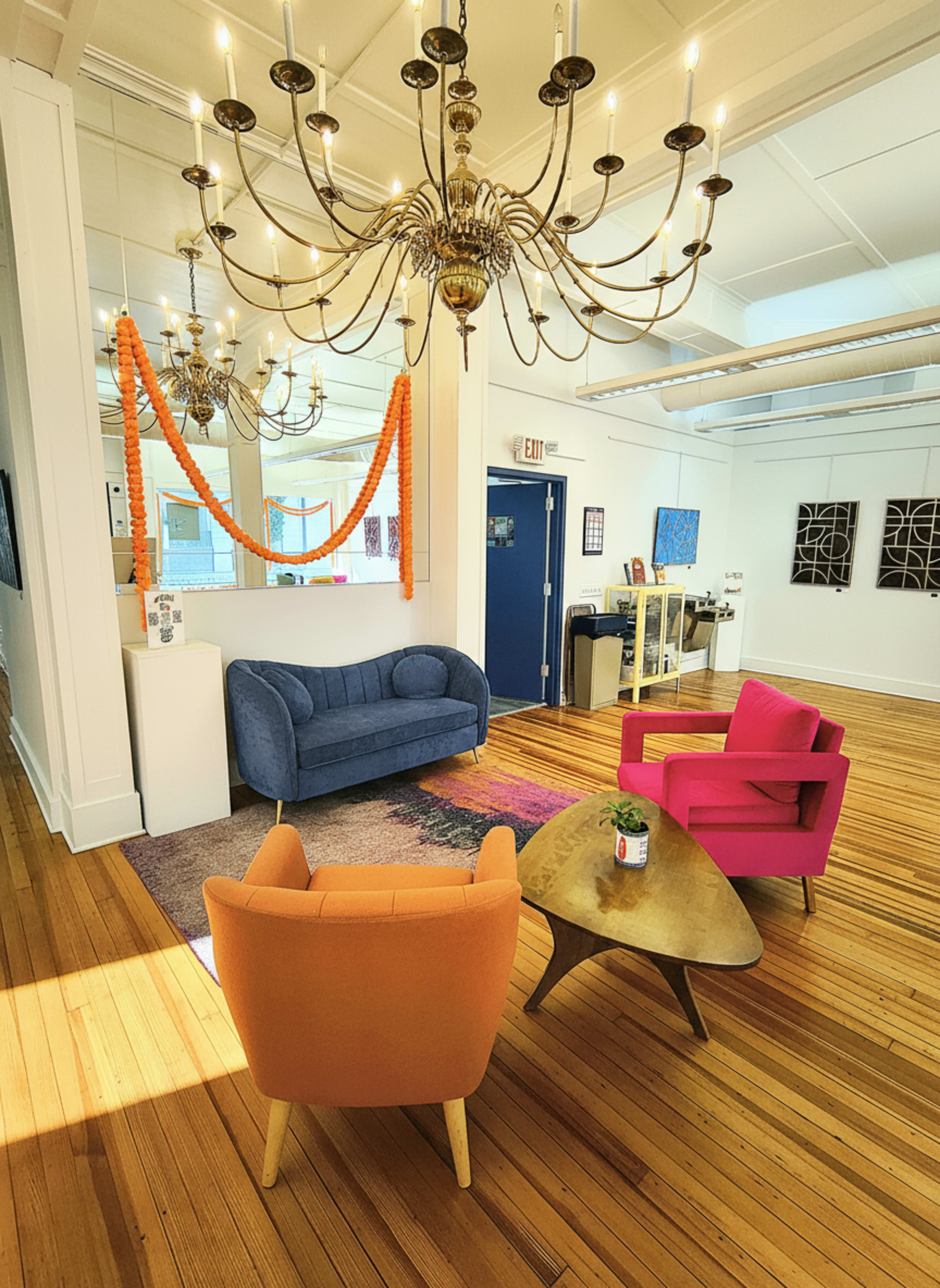 The image shows a modern lounge area with a blue sofa, two colorful chairs in orange and pink, a round table, and a chandelier, all set against a backdrop of wooden flooring and bright lighting.