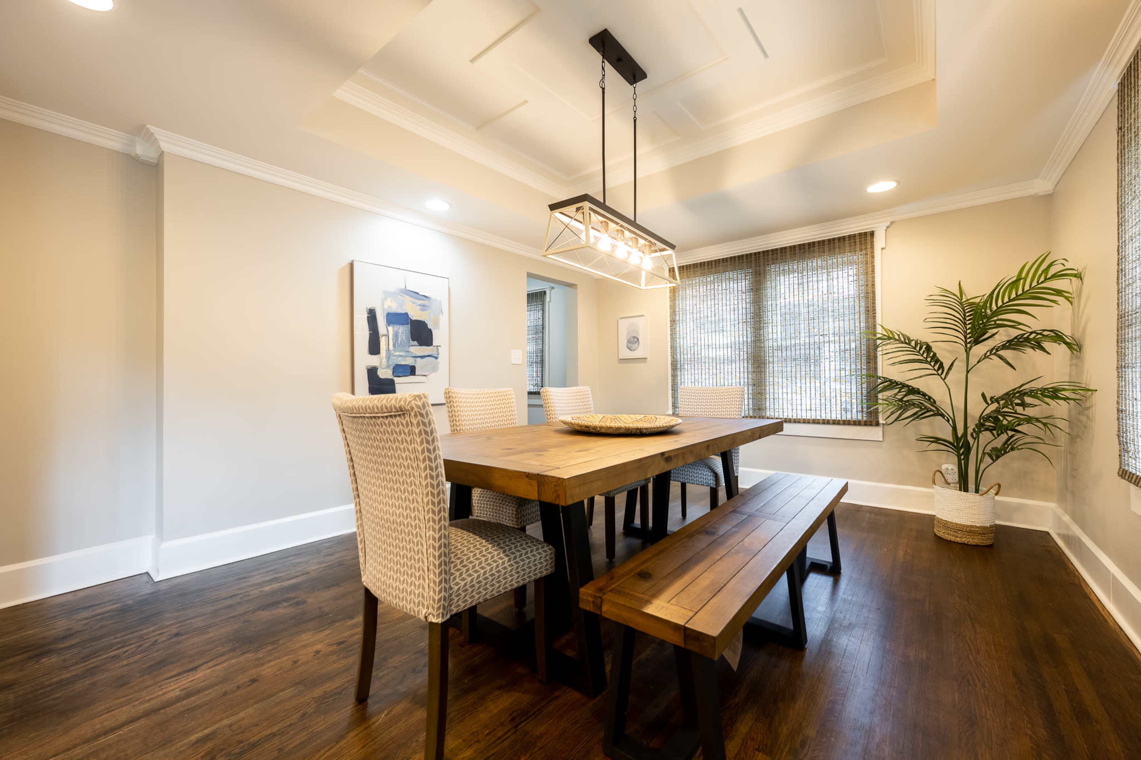 A wooden dining table with benches and chairs is set beneath a pendant light in a well-lit dining room featuring a potted plant and patterned window shades.