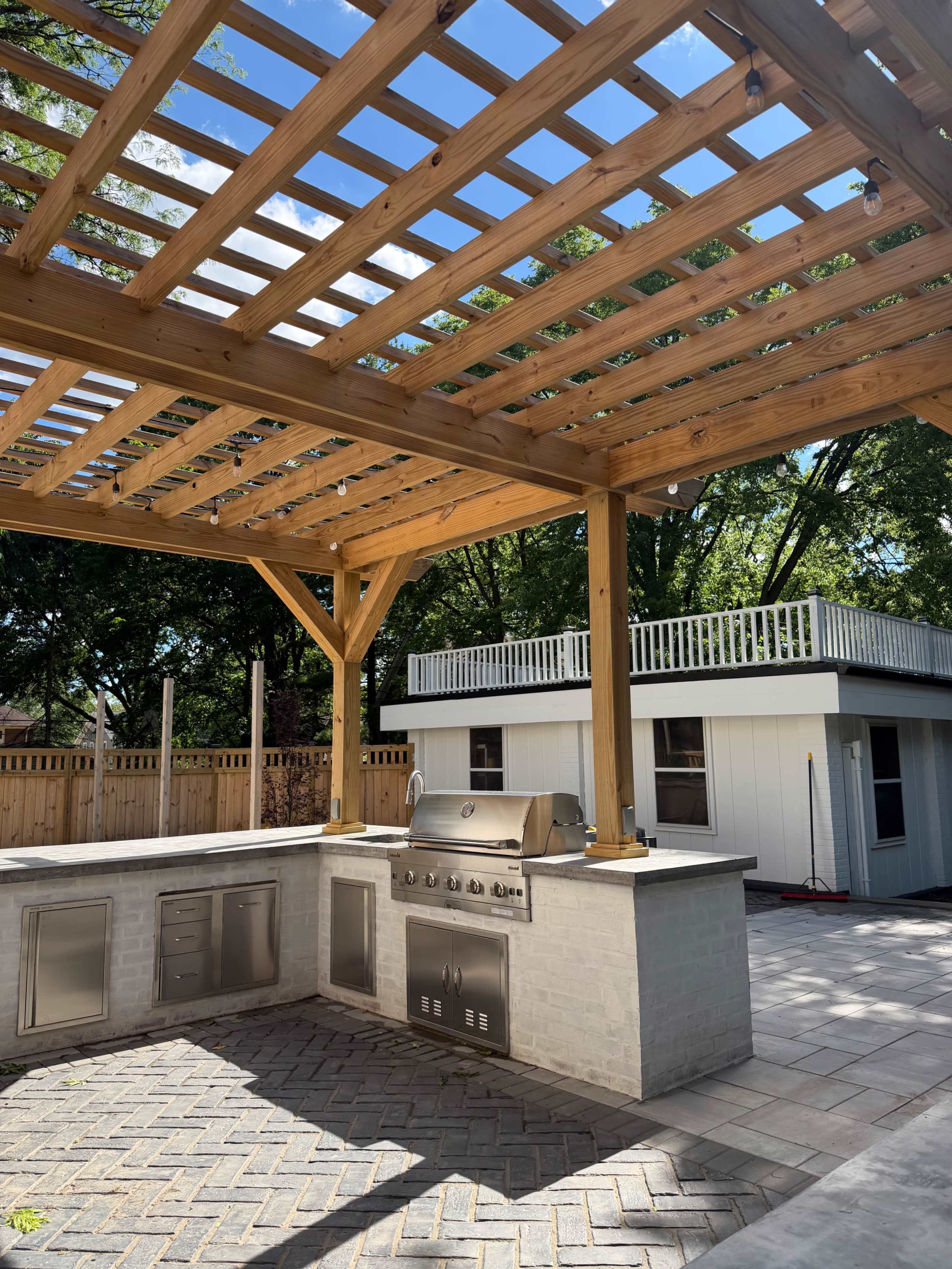 The image shows an outdoor kitchen area with a wooden pergola, featuring a grill and countertop, next to a house with a white exterior and a second-story balcony.