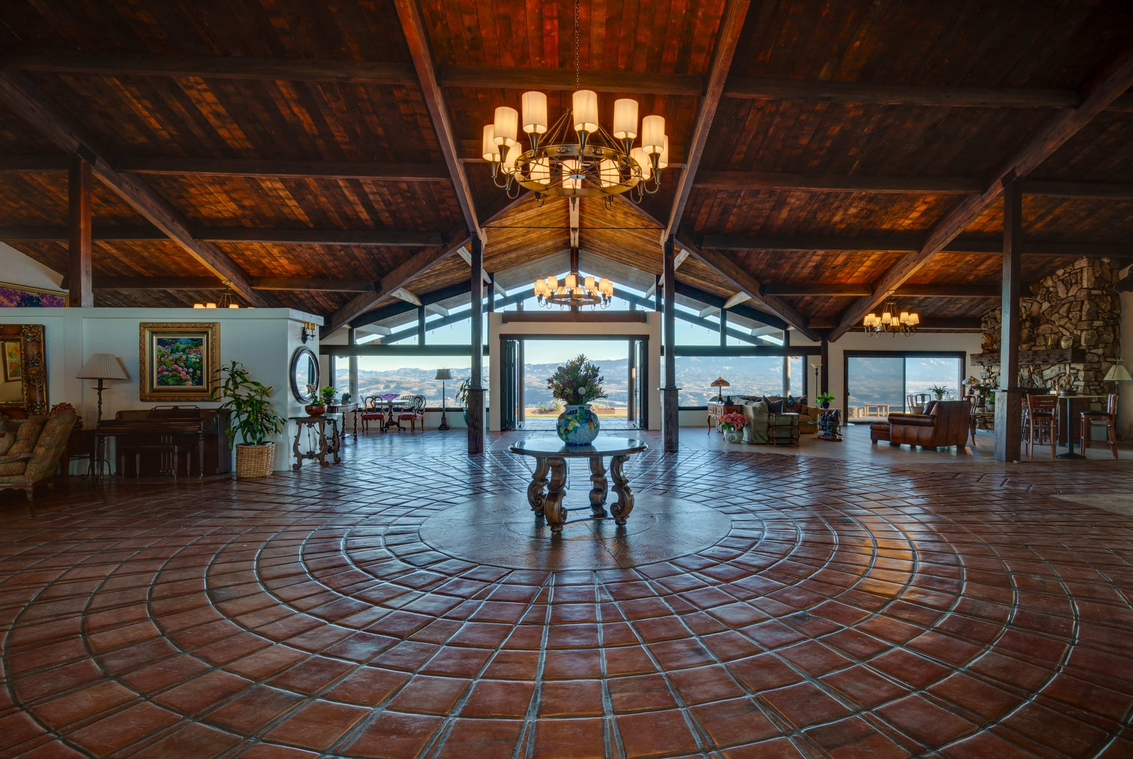 The image shows a spacious interior with high wooden ceilings, a tiled floor featuring a circular design, and large windows that offer a view of the outdoors.