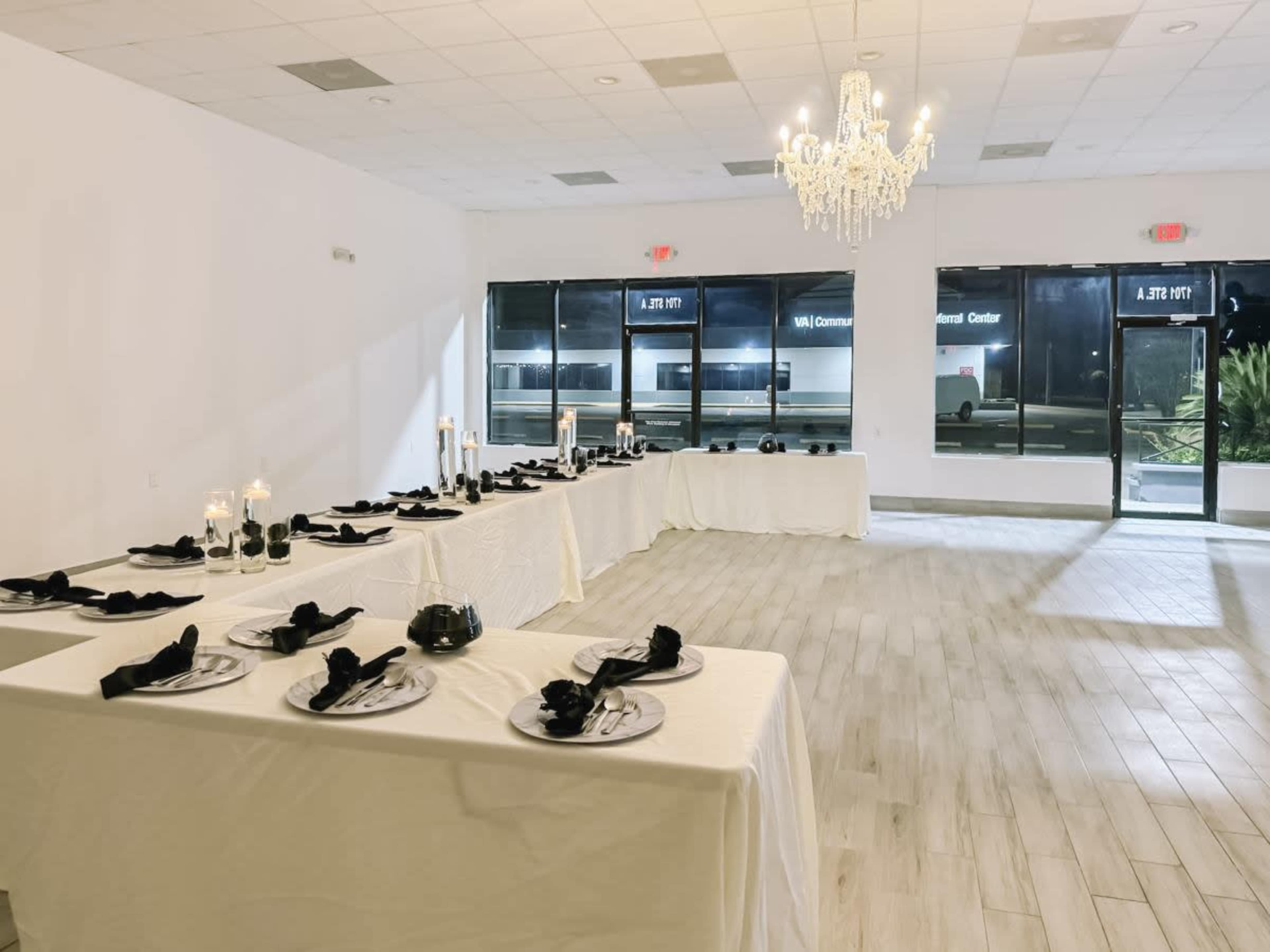 The image shows a spacious event room set with long tables covered in white linens, black napkins, and candlelit centerpieces, illuminated by a chandelier.