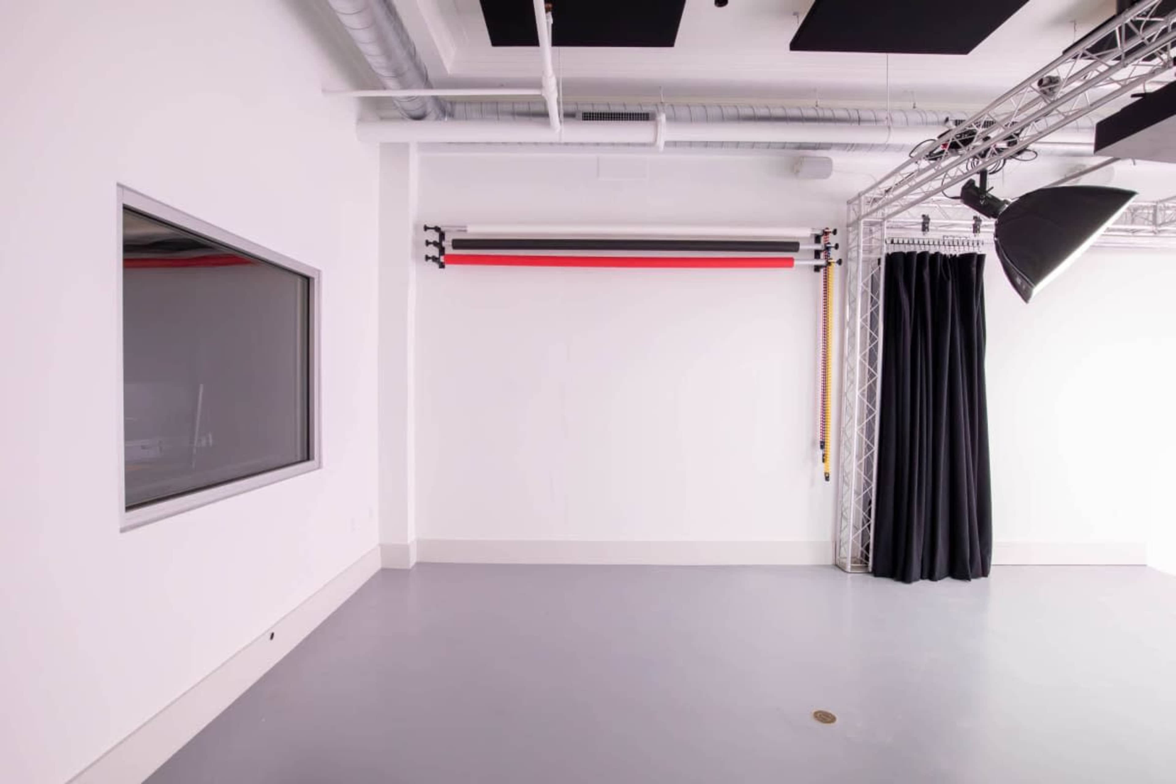 The image shows a minimalist studio space with white walls, a gray floor, a large window, a backdrop support system with colored rolls, and black curtains.