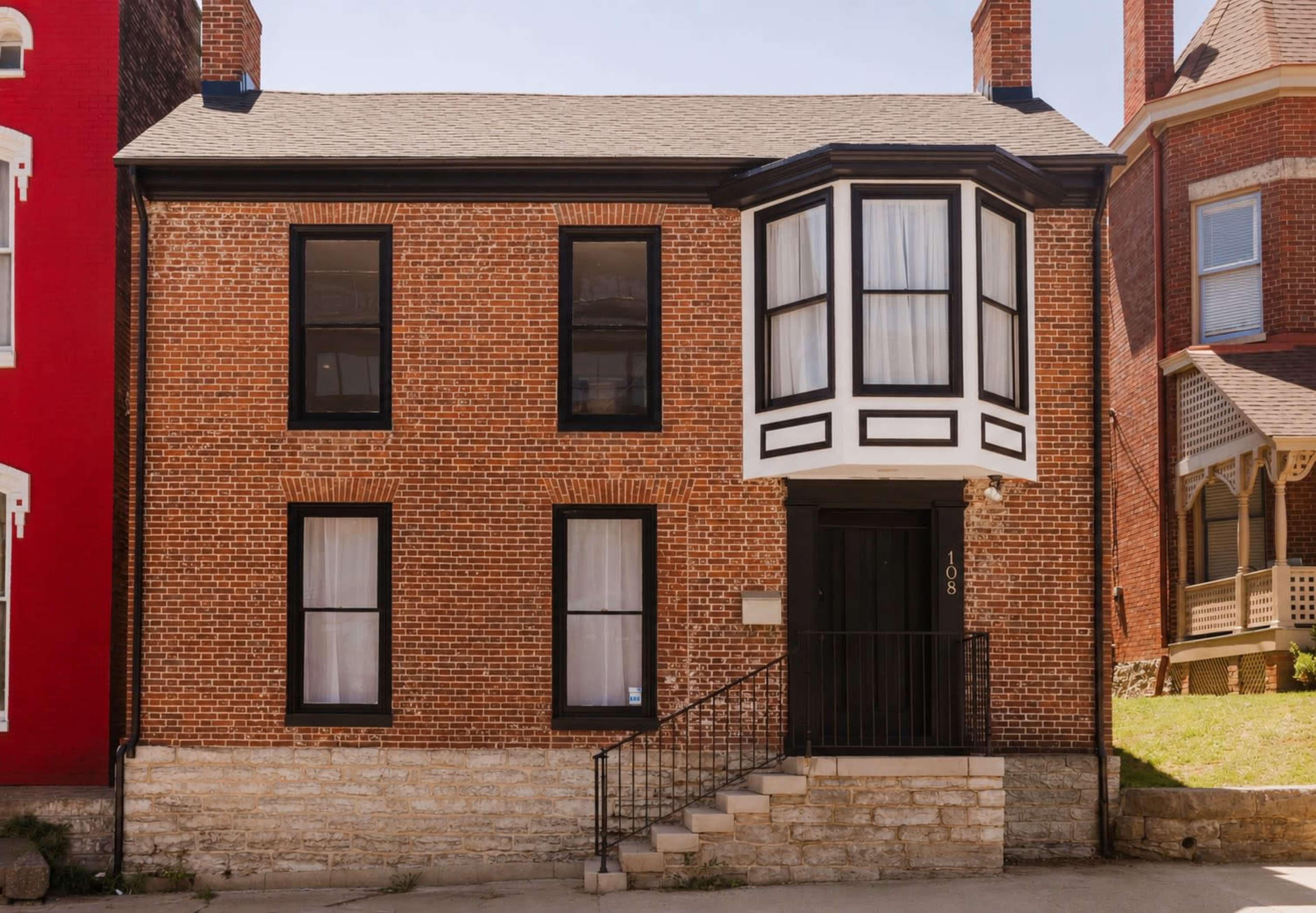 The image shows a brick building with a modern bay window and a set of stairs leading to the front door, flanked by two neighboring structures.