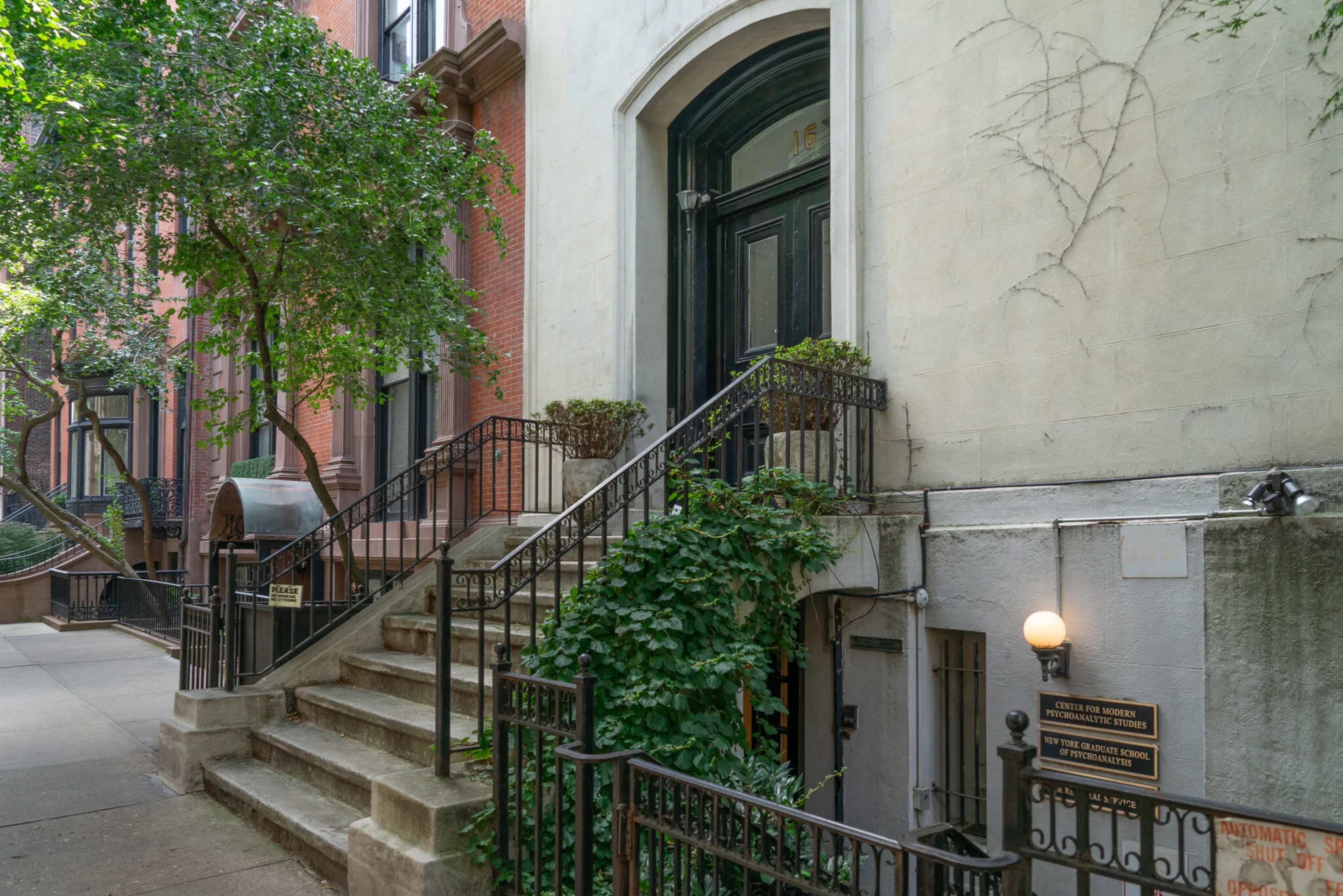 The image shows a brownstone building with a black door and staircase, surrounded by greenery and a wrought iron fence.