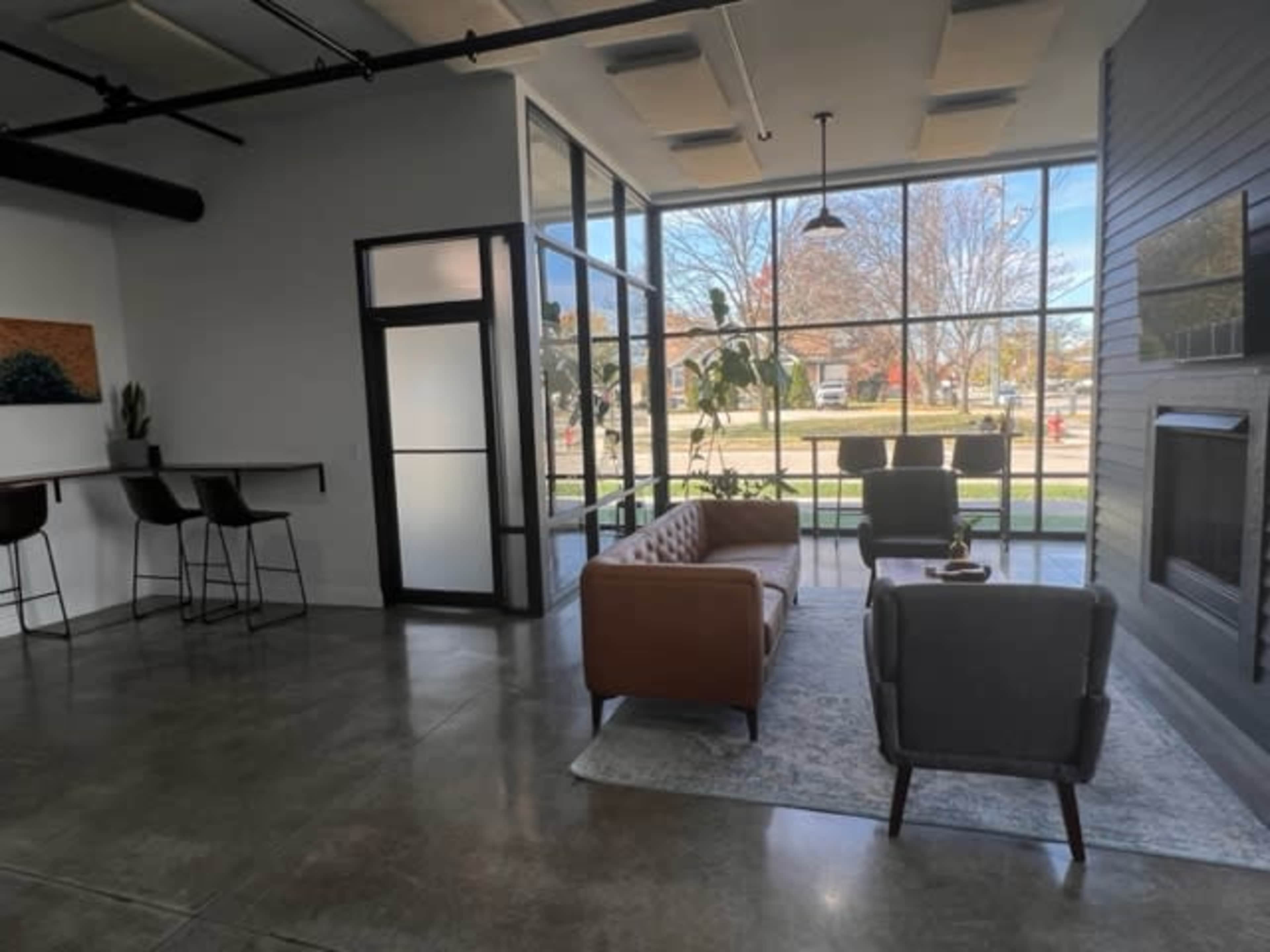 A modern waiting area with a brown leather sofa, a gray armchair, large windows, and a small bar with high stools.