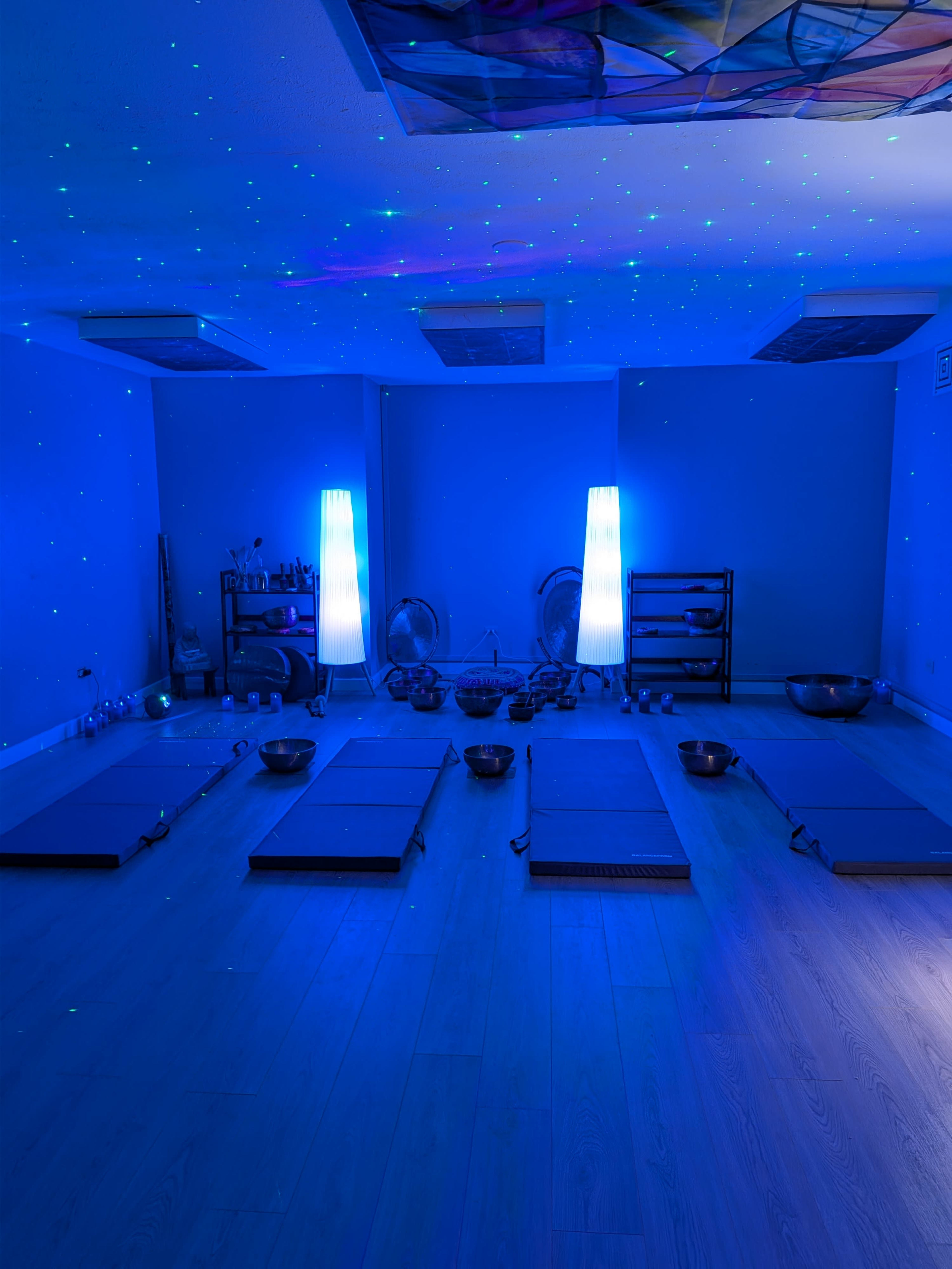 The room is set up for a sound therapy session, featuring six mats arranged on a wooden floor with bowls and soft lighting.