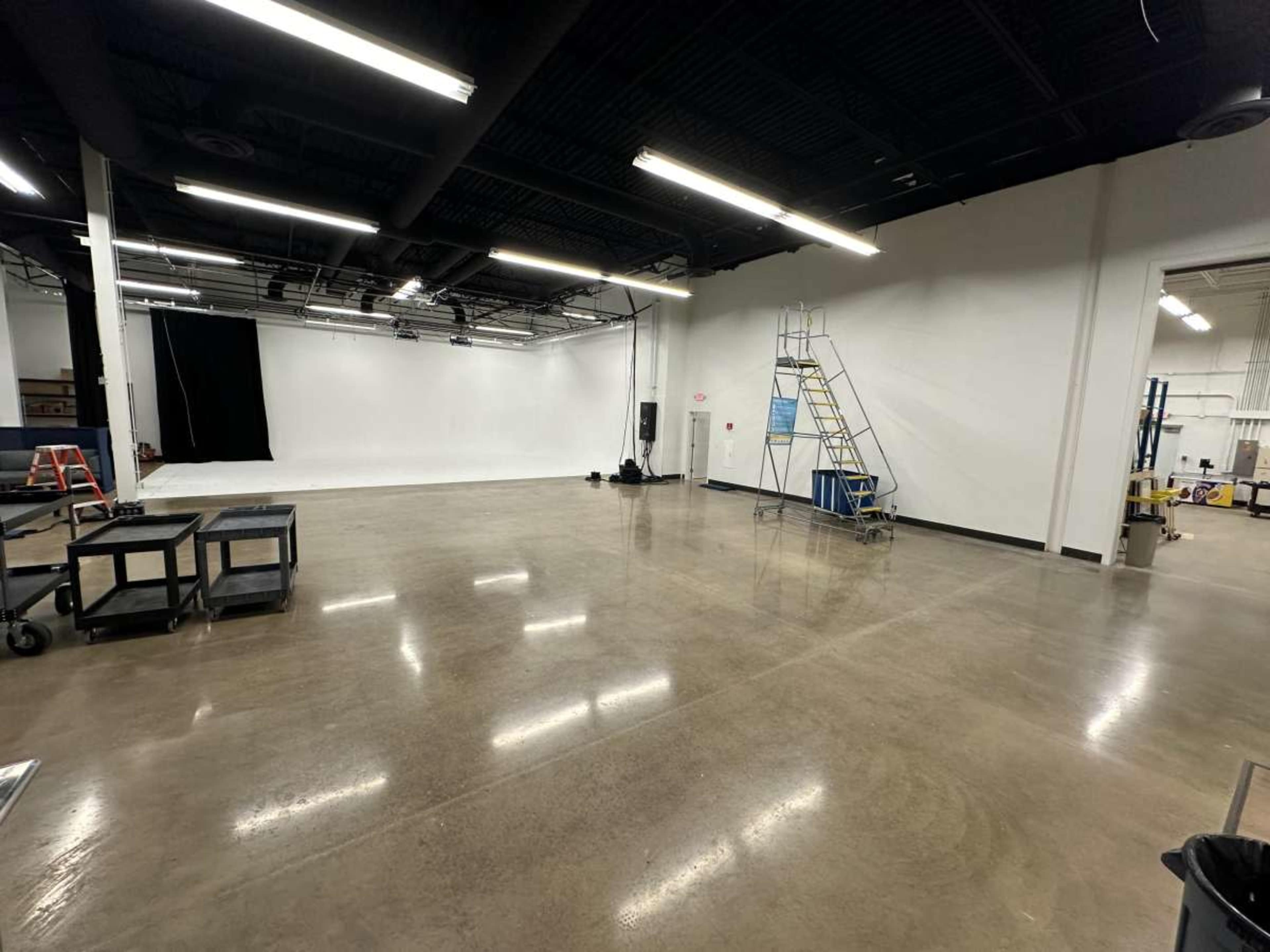 A spacious, empty studio with a polished concrete floor, white walls, and a black backdrop, along with a ladder and various equipment carts.